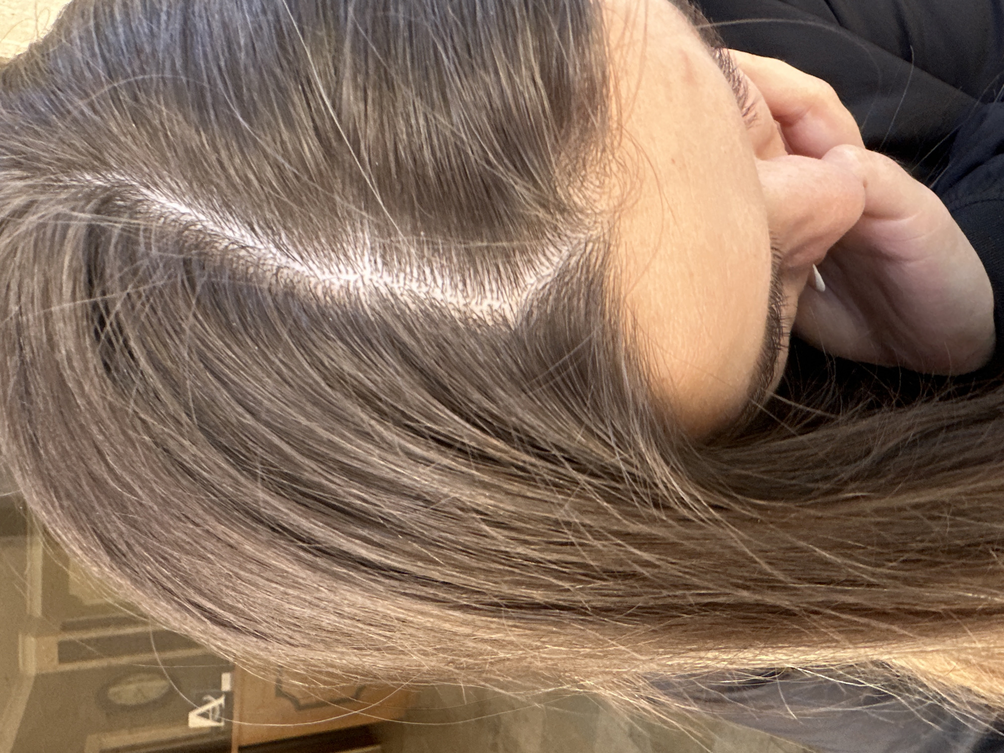The image shows a close-up view of a person's scalp and hair, with a distinct part visible. The hair is dark brown with some lighter strands. The background is blurred, but appears to be an indoor setting with furniture and architectural details that might suggest a historical building. There are no clear indicators of the city of Sozopol, Bulgaria. The focus is entirely on the hair and scalp.
