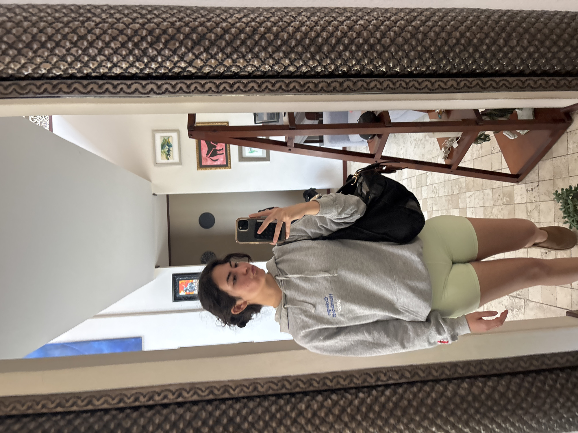 A person is seen taking a selfie in a mirror. The person is wearing a gray hoodie and light green shorts, with a black backpack slung over their shoulder. They are holding a phone up to their face, presumably to take the picture. The location appears to be inside a building in Puebla, Mexico, indicated by the architecture and style of decoration visible in the background, including framed artwork on the walls and patterned flooring. The setting includes furniture visible in the mirror reflection.