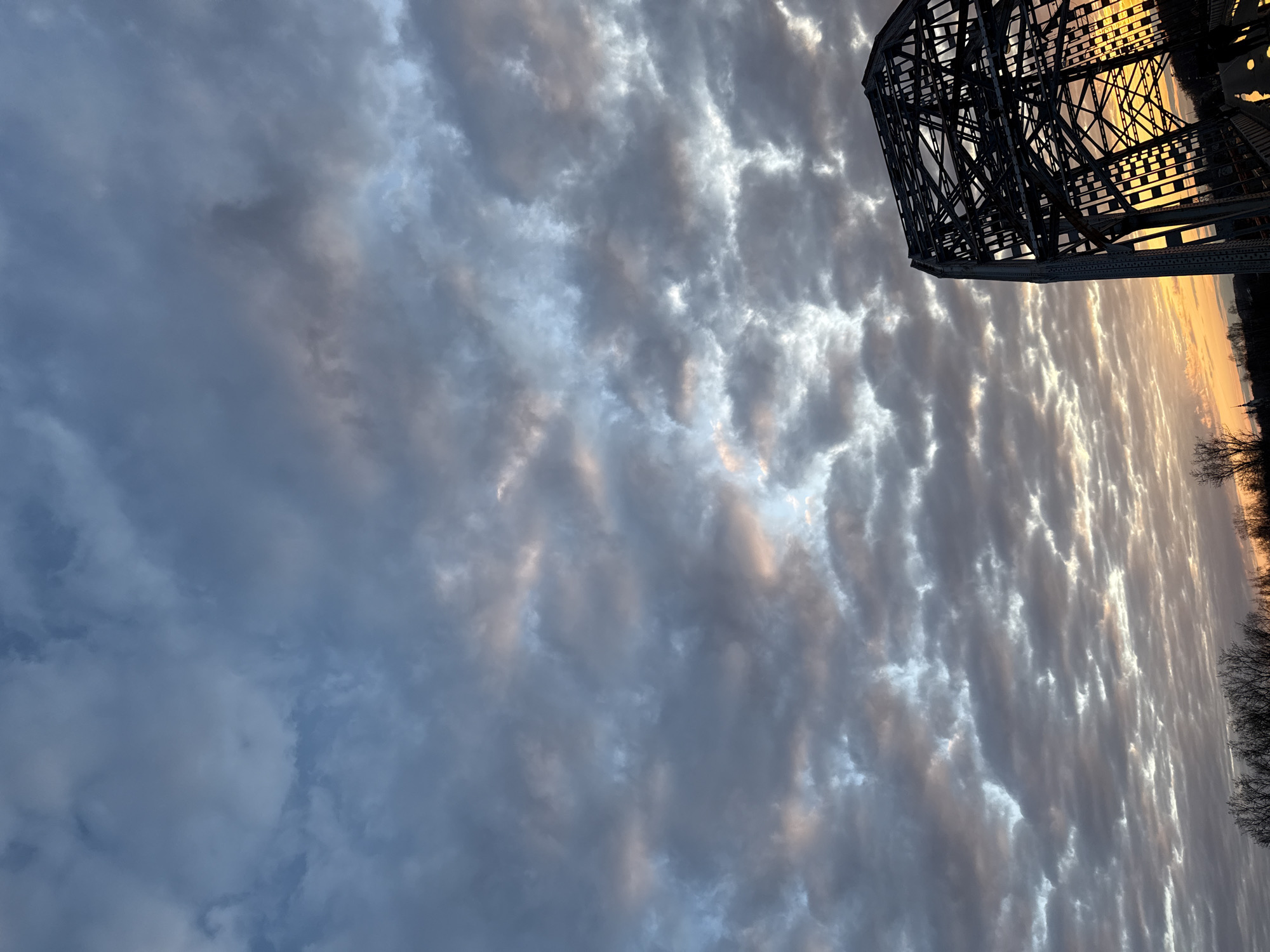This rotated image captures a dramatic cloudy sky and a prominent metal bridge, likely at either sunrise or sunset, over Dmitrov, Russia. The majority of the frame is dominated by a vast expanse of clouds, showcasing intricate patterns and textures. These clouds vary in color from deep blue-grey on the left to lighter tones of grey, white, and hints of soft pink and orange, particularly where they are illuminated by the sun.
On the right side of the image, a large, dark metal truss bridge is partially visible, its complex structural framework sharply silhouetted against a brilliant, golden-orange sky. The low sun's light streams through the bridge's crisscrossing beams, creating a striking contrast between the man-made structure and the radiant sky. A faint, dark line of trees or land can be seen along the very bottom right edge, further silhouetted against the bright horizon. The overall lighting suggests a specific time of day when the sun is very low, casting a warm glow and creating strong atmospheric effects on the clouds. There are no people visible, nor any discernible text in the image. The scene evokes a sense of tranquil grandeur, highlighting the interplay of natural light and an engineered landmark.