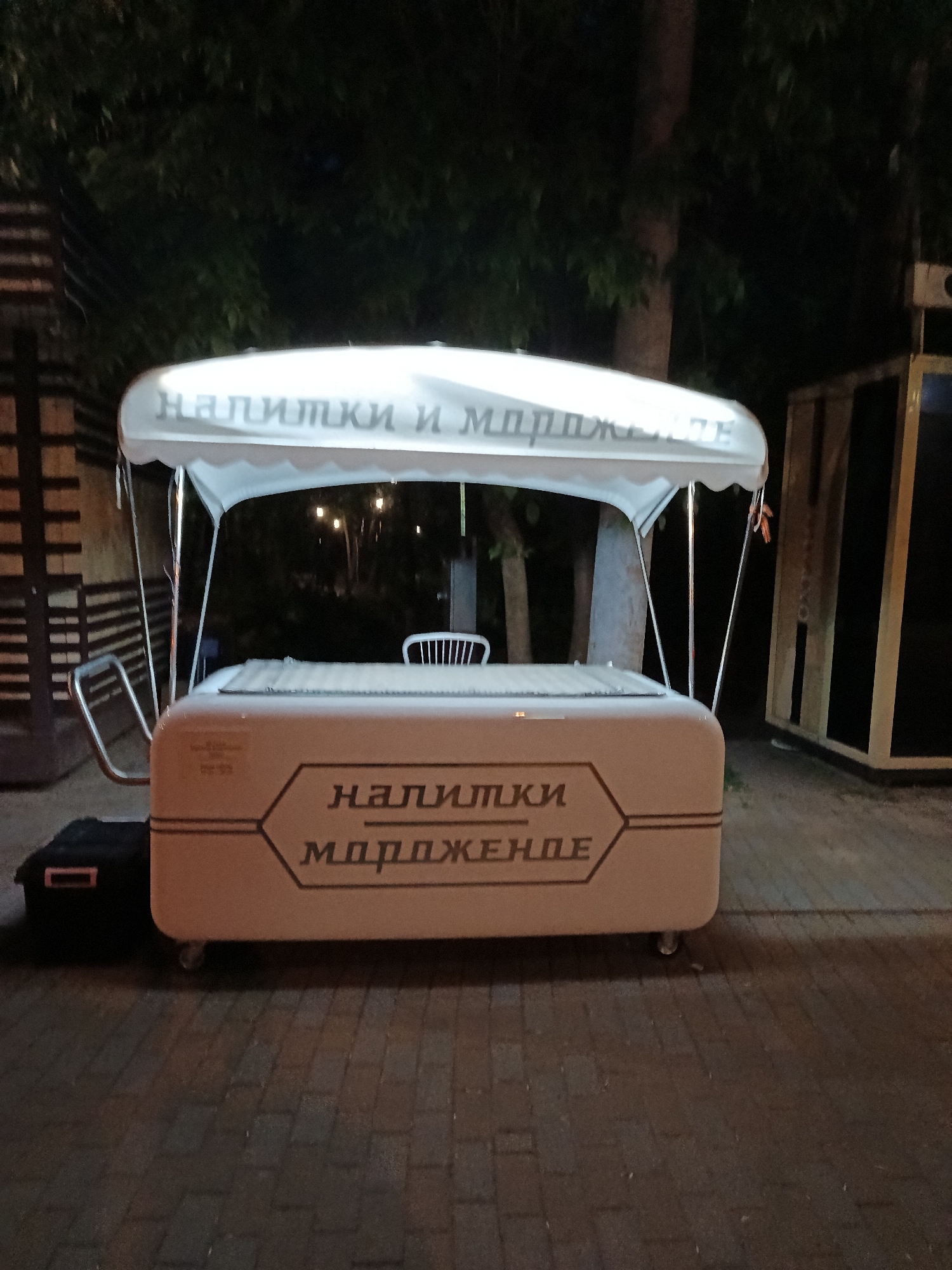 The image captures a mobile vendor cart set up outdoors at night or late evening in Moscow, Russia. The central subject is a light-colored cart with a white canopy, equipped with wheels, suggesting portability. The canopy features the Cyrillic text "НАПИТКИ И МОРОЖЕНОЕ" (Drinks and Ice Cream), indicating the cart's purpose. The front panel of the cart reiterates this, displaying "НАПИТКИ" (Drinks) above "МОРОЖЕНОЕ" (Ice Cream) within a stylized, angular border.
No people are visible in the scene. The cart appears to be illuminated by an internal light source, contrasting with the dark background. Behind the cart, a white chair is faintly visible, and in the distance, some sparse artificial lights can be seen. The background consists of dark foliage and tree trunks, suggesting a park or green area. To the left of the cart, a wooden, fence-like structure is partially visible. To the right, a dark, box-like structure with light-colored framing stands on the ground. The ground beneath the cart is paved with what appears to be dark cobblestones or bricks. A black storage box or container rests on the ground to the left of the cart. The cart is stationary, likely set up for business or closed for the night, offering drinks and ice cream.