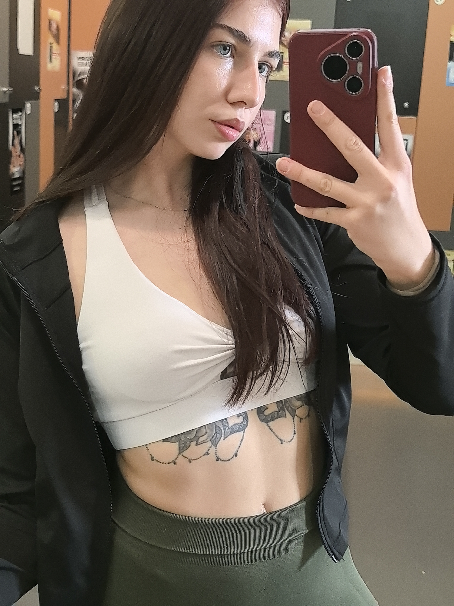 A woman is holding up a burgundy smartphone in her right hand to take a selfie. She has dark brown hair and light blue eyes. She's wearing a white sports bra, a black jacket, and olive-green leggings. A floral tattoo is visible on her lower abdomen. The background is a locker room setting with lockers. It's unlikely that the specific location is in Stara Zagora, Bulgaria.