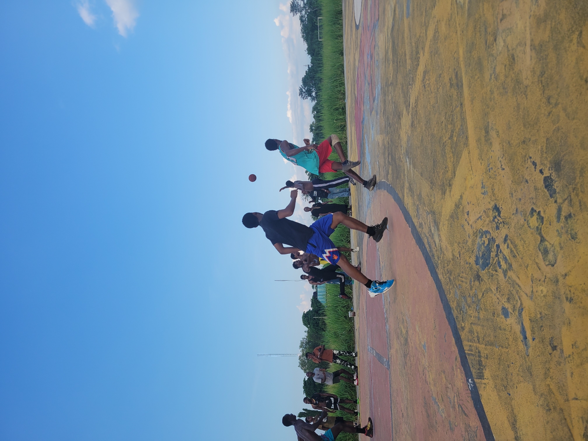 A group of young men are playing basketball on an outdoor court. Two players in the foreground are in motion, one dribbling and the other running. Other players are visible in the background, watching the game. The court is worn and painted with colorful markings, and tall grass and trees line the perimeter. The sky above is a clear blue with a few white clouds. The scene appears to be in a rural or semi-urban area, possibly in Egbagu, Nigeria, given the context.