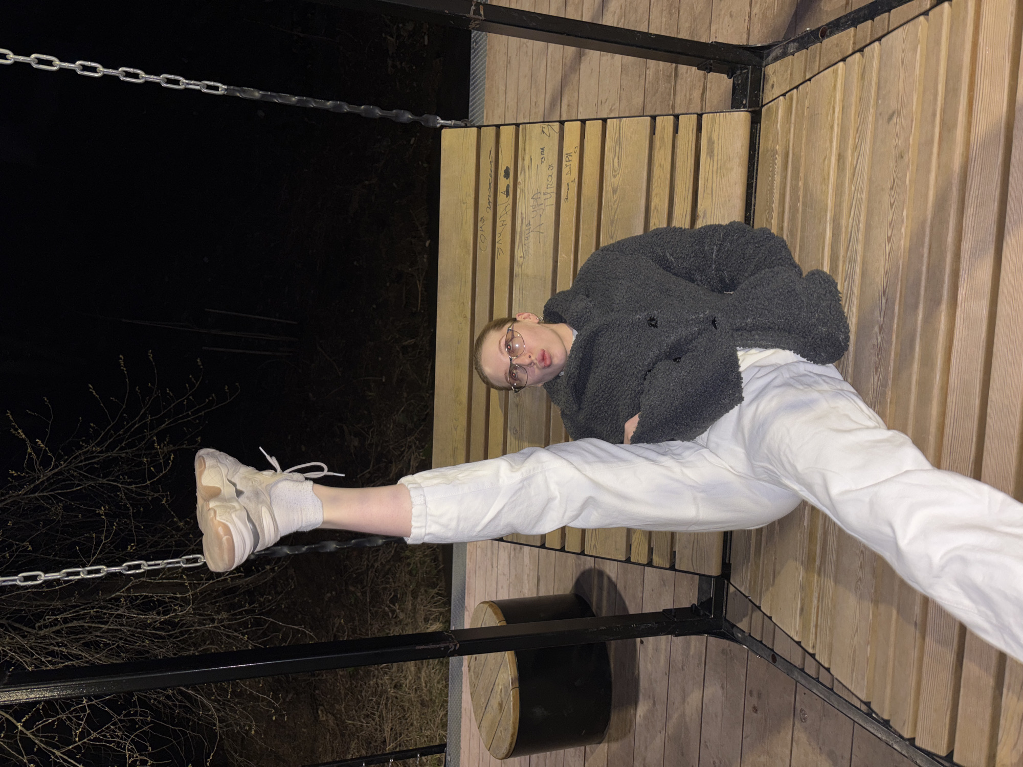 An individual, likely female, with short light hair and glasses, is captured in a playful pose on an outdoor wooden platform at night. She is dressed in a dark, fluffy jacket, white pants, and light-colored sneakers with white socks. Her lips are pursed, giving her a pouting or whimsical expression. She is lying on the wooden slats, with one leg extended upwards and the other bent.
The setting is an outdoor space, likely a park or public recreation area in Perm, Russia, characterized by a sturdy wooden platform or bench structure with vertical wooden slats and black metal framing. A metal chain runs horizontally across the left side of the frame, near her outstretched leg, and a dark, cylindrical, log-like object serves as a seat or decorative element below her. The background is dark, revealing bare tree branches against the night sky, illuminated only by artificial light from above, which casts soft shadows.
Notable details include various inscriptions and graffiti carved or written in Cyrillic script on the wooden slats behind the person's head. Legible phrases include "Сонч" (Sonch, likely a nickname), "злая чёлка" (evil fringe/bang), "А-ДУРА" (A-FOOL/STUPID), "Ксюша" (Ksyusha, a common name), and "Паша" (Pasha, another common name). Other less clear writings are also present, such as "тзпа" and "Димка лура." The overall mood is casual and slightly artistic, indicative of an impromptu photo opportunity.