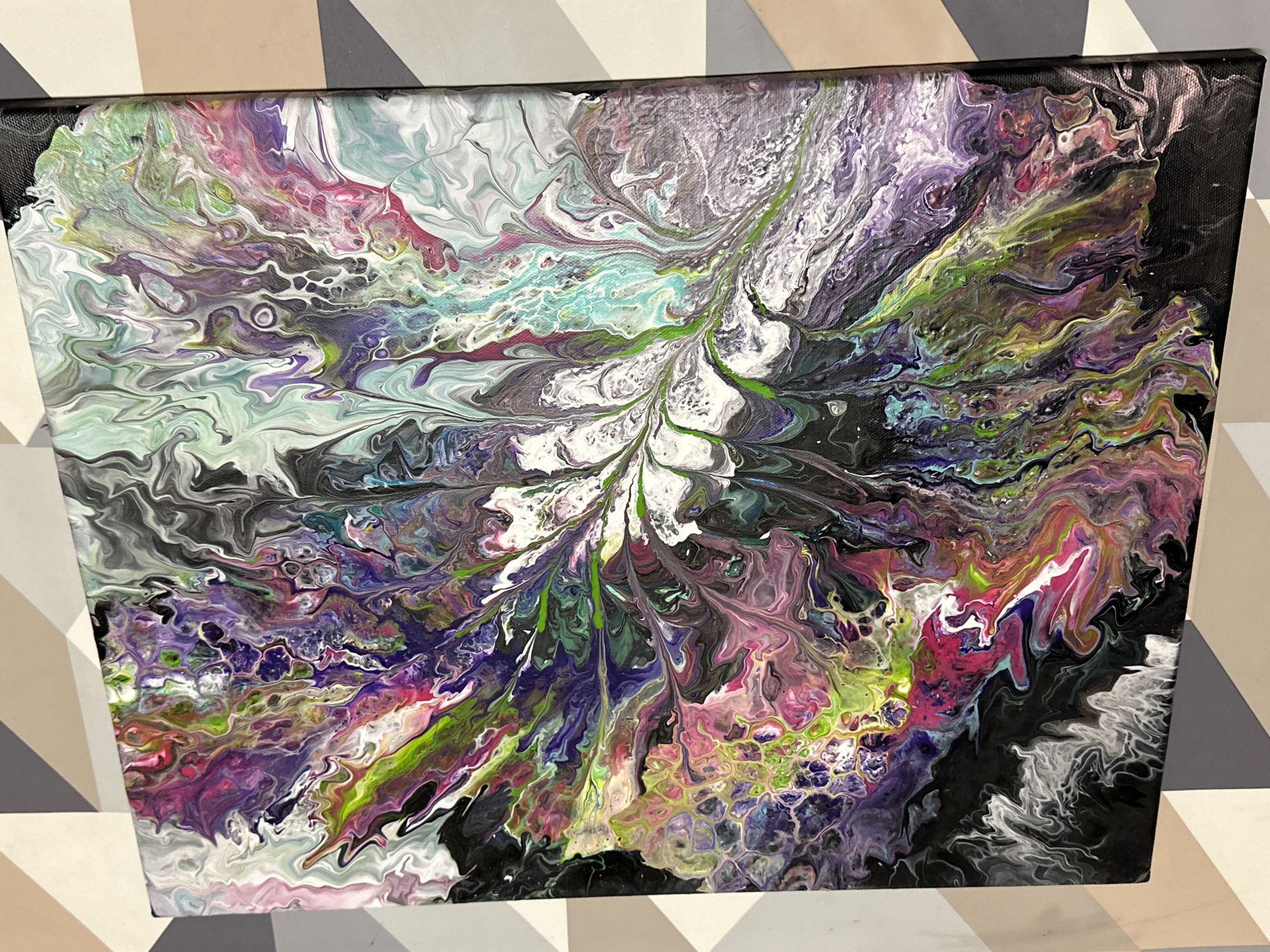 The image is a close-up, tilted shot of an abstract acrylic pour painting on a canvas. The painting features a vibrant and chaotic explosion of colors, dominated by shades of purple, pink, green, turquoise, black, and white. The paint has been poured and manipulated to create organic, swirling patterns with a fluid, marbled effect. Some areas show cell-like formations, while others exhibit a feathery or dendritic structure. The canvas is framed by a black edge, and the painting is displayed against a background of geometric wallpaper in beige, gray, and off-white diagonal patterns. The lighting casts shadows, highlighting the textured surface of the paint. There are no people, animals, or any recognizable objects from the city of Villa Pasquali, Italy, present in the image.