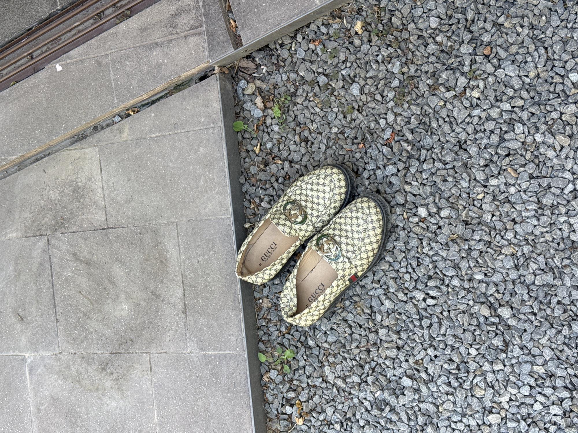The image depicts a pair of Gucci loafers lying on a bed of gray gravel. The gravel appears to be part of a landscaping feature adjacent to a stone-tiled walkway or patio. A small amount of vegetation, including a few scattered blades of grass and a small weed, is visible among the gravel. The location is indicated as Castel Gandolfo, Italy, but there are no obvious landmarks or buildings visible in the image that can be used to confirm this. There are no people present in the image.
