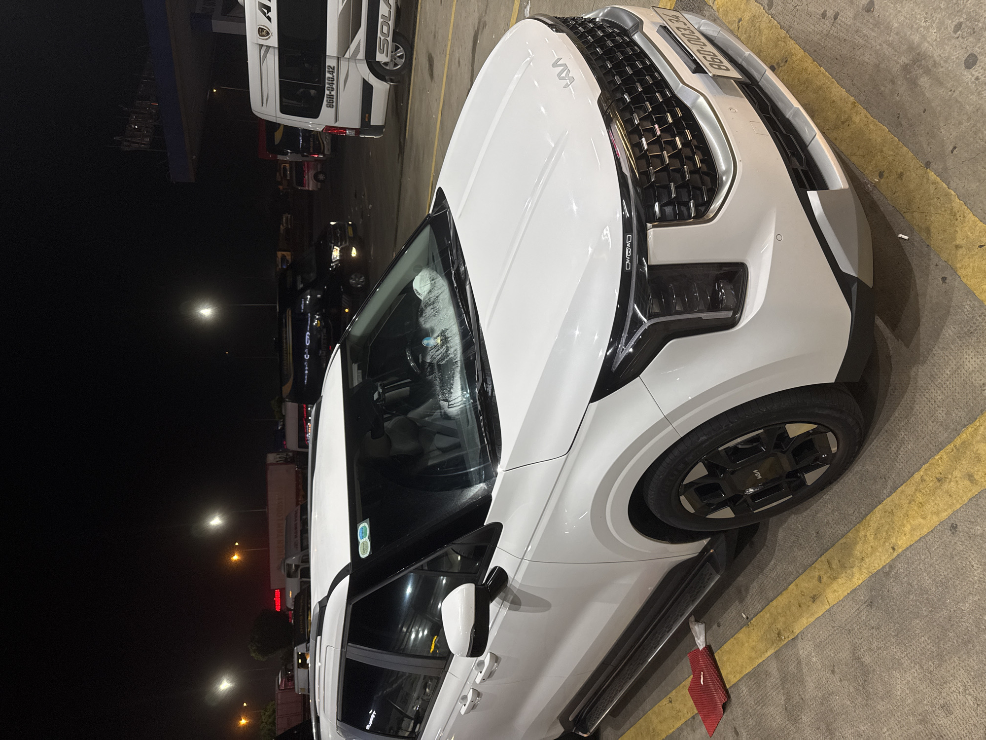 A white Kia Carnival minivan is parked at a gas station at night. The minivan is positioned at an angle, facing slightly to the right, with its front prominently displayed. The gas station has yellow lines on the ground indicating parking spots, and a red mat is placed on the ground near the front left wheel. In the background, other vehicles are visible, including a white bus with "SOLAR" written on its side, and a black van. Several streetlights are illuminated, casting a glow on the scene. The overall environment suggests a roadside rest stop or a gas station in a rural or suburban area of Ấp Xuân Tiến, Vietnam. There are no people visible in the image.