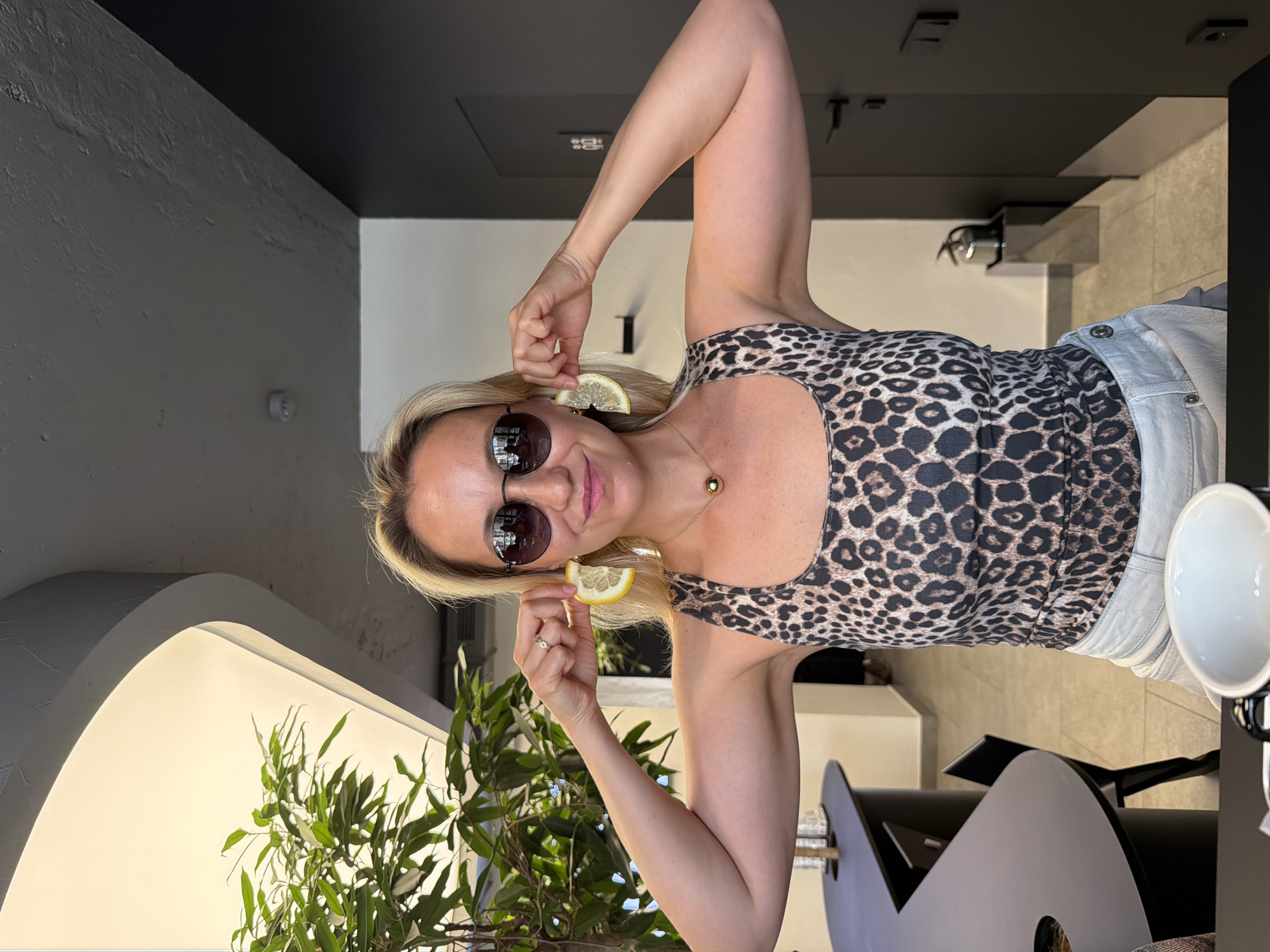 A blonde woman wearing sunglasses and a leopard-print tank top holds lemon slices up to her ears as earrings. She is standing in a modern cafe, with a large green plant to her left and a dark gray door to her right. The cafe has a gray ceiling and light-colored tile flooring. The woman has a gold ball necklace on and is wearing a ring on her right index finger.