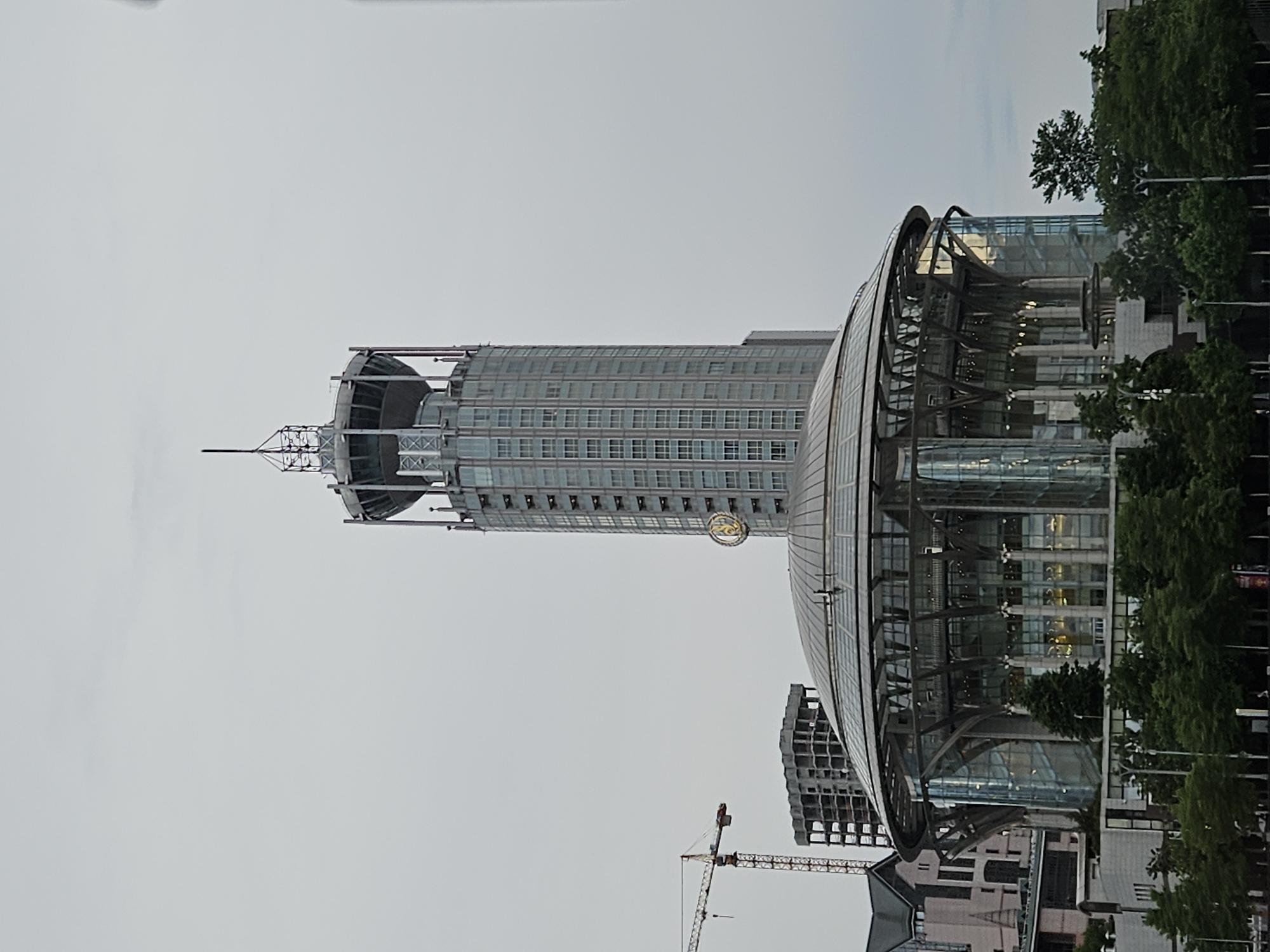 The image depicts a scene in Moscow, Russia, featuring the Swissôtel Krasnye Holmy Moscow hotel and the Moscow International House of Music. The tall hotel tower is visible on the left, topped with a distinctive antenna-like structure. A round glass structure, the International House of Music, sits to the right. In the foreground, there are trees and other buildings. A construction crane is visible in the distance. The sky is overcast.