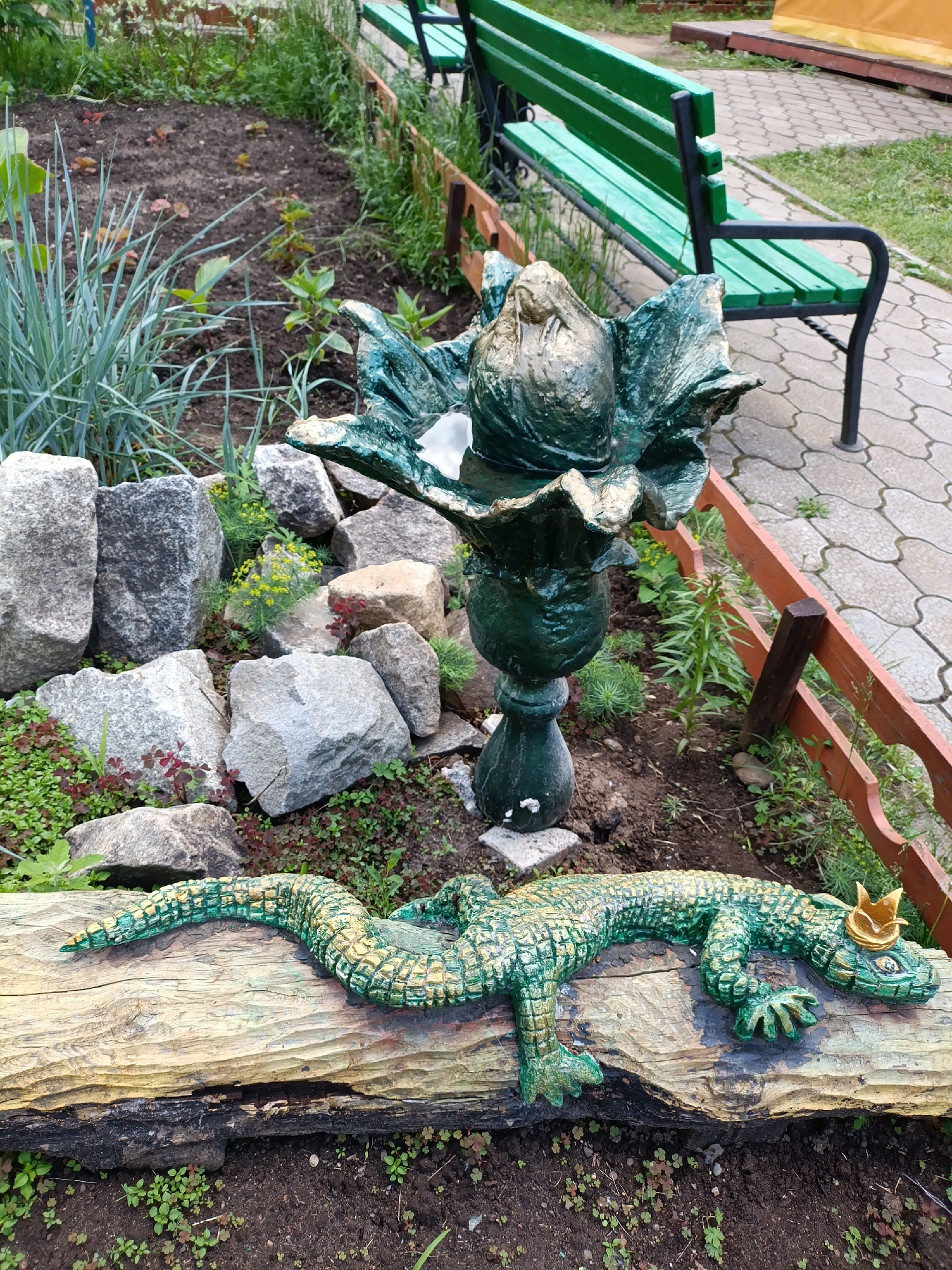This image captures a vibrant and decorated garden area in Chaikovsky, Russia. The scene features a landscaped bed bordered by a low reddish-brown wooden fence and an arrangement of various-sized rocks in shades of grey, white, and brown.
At the center of the garden bed stands a prominent green and gold sculpture resembling a stylized flower or plant, elevated on a matching pedestal. Closer to the foreground, a charming green and gold lizard sculpture rests on a large, weathered wooden log. The lizard has intricate scale patterns and wears a small, golden crown on its head, adding a whimsical touch to the decor.
The garden bed itself is populated with various plants, including tall, spiky green foliage, smaller ground cover with green and reddish leaves, and patches of small yellow flowering plants nestled among the rocks. The soil appears dark and well-tended.
To the right of the garden, a grey interlocking paved pathway is visible, on which a green park bench with a black metal frame is situated. The light is bright and natural, suggesting daytime conditions, and there are no harsh shadows. No people are present, and no text is visible in the image. The overall atmosphere is peaceful and decorative.