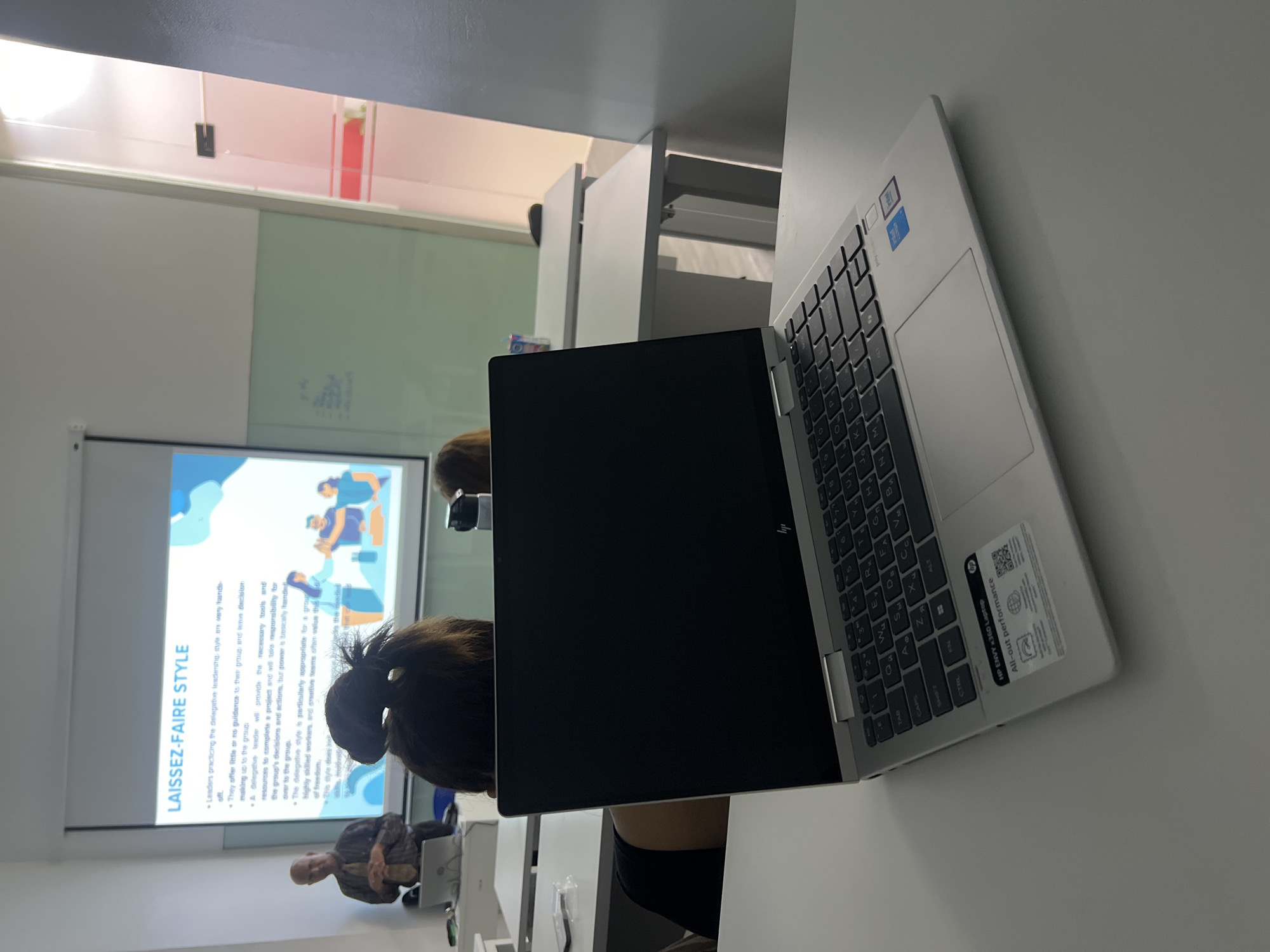 In a modern classroom setting, a laptop is positioned on a white table in the foreground, angled towards the viewer. The laptop's screen is black, and its keyboard is visible, displaying a QWERTY layout with Cyrillic characters. Stickers indicating "HP ENVY x360 Laptop" and "All-out performance" are affixed to the laptop's body. In the background, a projector screen displays text related to "LAISSEZ-FAIRE STYLE" with bullet points describing leadership characteristics. A presenter, a man with short hair wearing a collared shirt and tie, stands to the left of the screen, facing the audience. Several students are seated at tables, with the back of a woman's head and dark hair prominently visible in the mid-ground. The room features white tables and chairs, a light green wall section, and a glass-partitioned area with hints of red in the background, suggesting it might be an office or meeting space within a larger building. The overall environment appears to be an educational or professional training session. This scene is located in a classroom at the Universiti Tunku Abdul Rahman (UTAR) in Kuala Lumpur, Malaysia.
