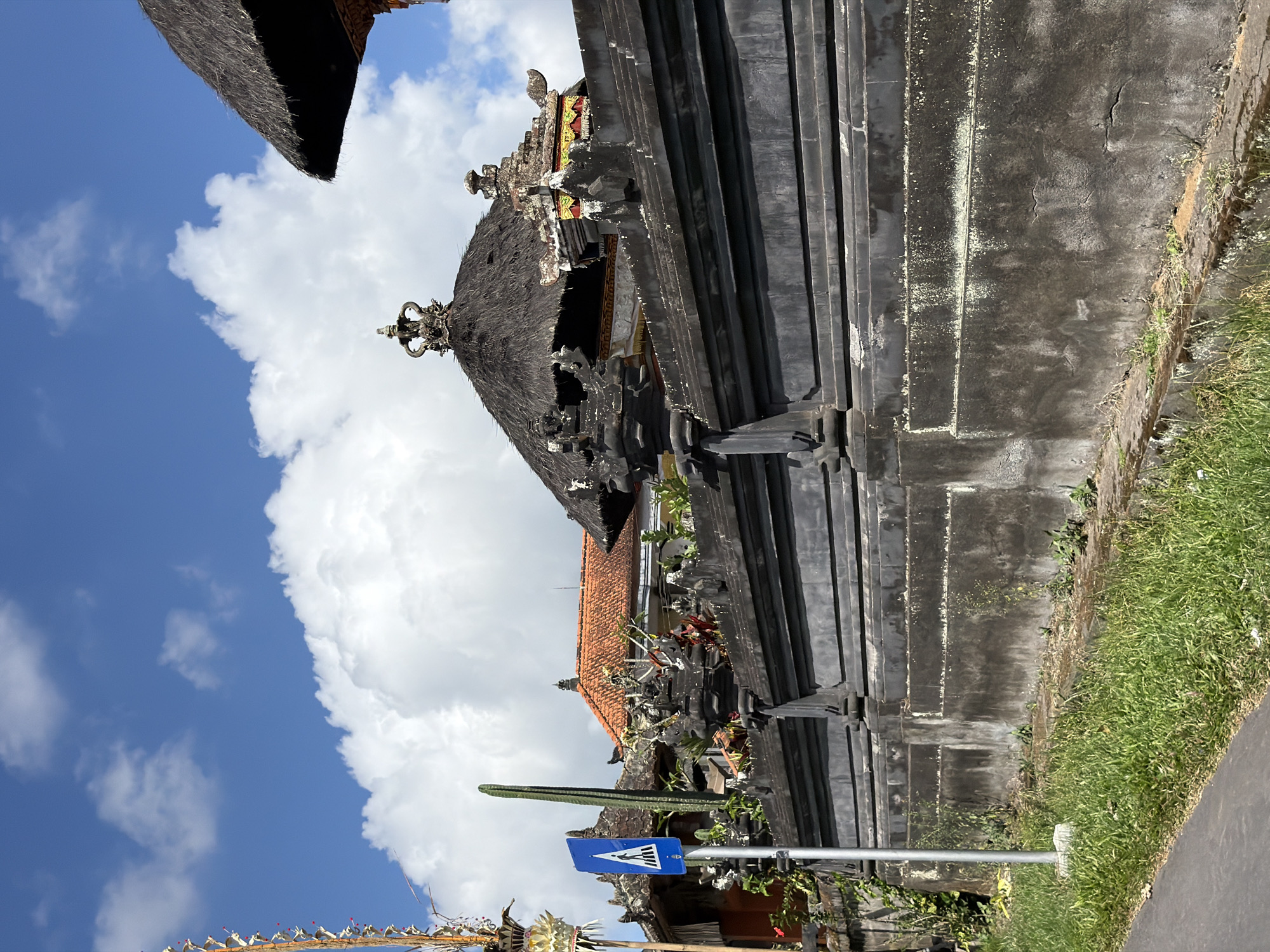 The image shows a pedestrian crossing sign on a metal pole in front of a traditional Balinese temple wall. The wall is made of dark stone and is decorated with intricate carvings. Behind the wall, parts of the temple buildings are visible, including thatched roofs and orange tiled roofs. The sky is blue with white clouds. The scene is located in Baturiti, Indonesia, which is known for its many temples and agricultural landscapes. There are no people or actions depicted in the image.