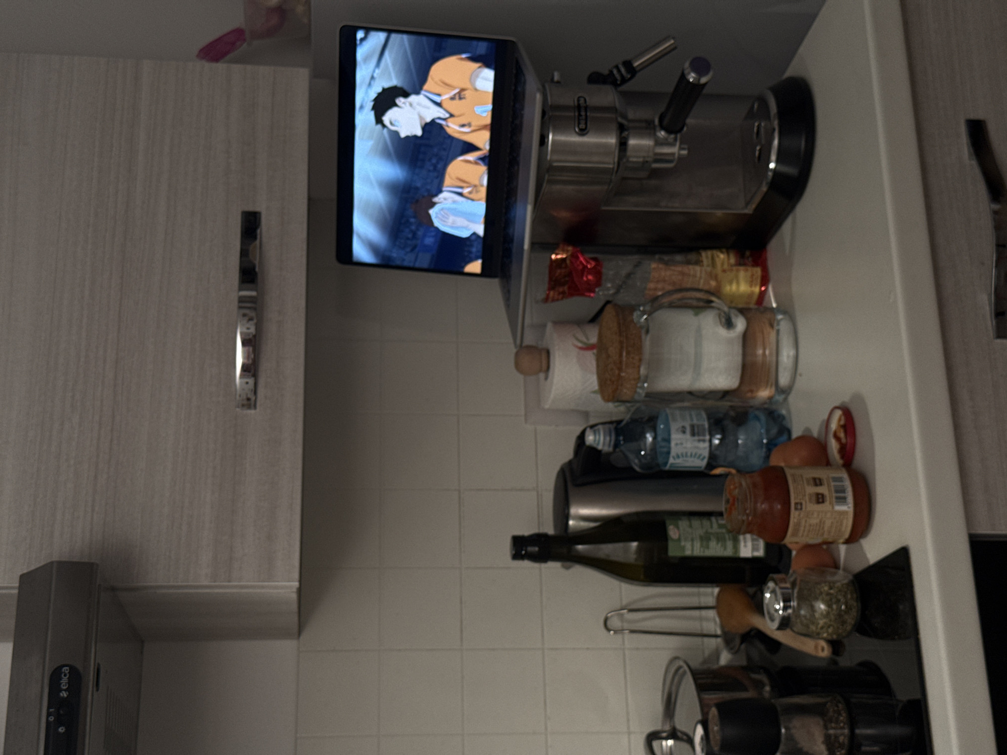 The image displays a kitchen counter area, rotated 90 degrees clockwise, revealing a functional and lived-in space.
On the left side of the upright image (top-left in the provided orientation), part of a light-colored wooden cabinet with a vertical, metallic handle is visible. Below it, a grey range hood with the "Elica" brand name printed vertically is mounted.
The central focus includes an open laptop resting on the counter. Its screen displays a vibrant anime scene featuring two male characters, possibly volleyball players, in sports uniforms against a blue background. Adjacent to the laptop, a silver espresso machine is positioned.
Further to the right (bottom-right in the provided orientation), the counter holds an array of kitchen items. These include a roll of paper towels, a clear plastic water bottle with some text visible on its label, and a dark green bottle, likely containing olive oil. Several jars are also present: one with a cork lid, a smaller glass jar filled with green herbs or spices, and a larger jar containing a reddish-orange sauce. The label on the sauce jar clearly shows the word "Passata" along with a barcode and other text. Beneath these, a wooden spoon, a spice grinder, and a pot with a lid are arranged, some resting on a metal rack.
The background wall is covered with light-colored square tiles. The lighting indicates an indoor setting, likely under artificial illumination. The scene suggests someone is engaged in kitchen activities, such as cooking or preparing food, while simultaneously watching content on the laptop. No specific time of day or emotions are discernible from the image.