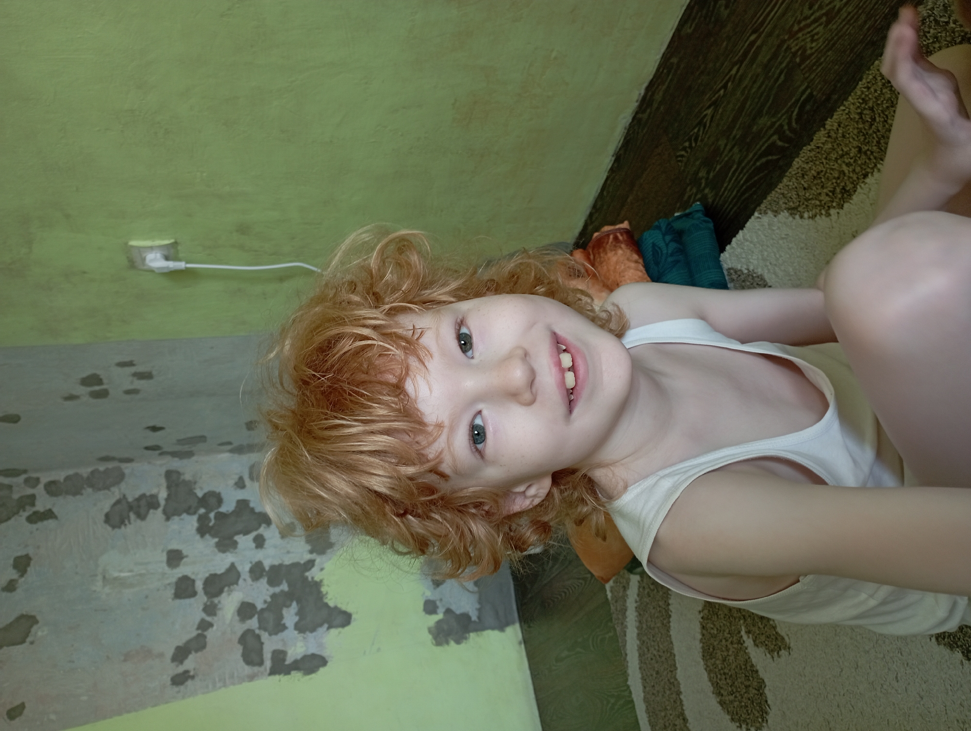 The media file shows a close-up, rotated image of a young child with red, curly hair lying on their side. The child is wearing a white tank top and appears to be smiling, with their mouth open slightly revealing their teeth. Their eyes are open and looking towards the camera. Freckles are visible on their cheeks and nose.
The background includes a textured green wall with some peeling paint or plaster, suggesting an indoor setting, possibly a room. To the right of the child, a patterned rug and dark wooden floor are visible. There are also some folded items of clothing or fabric in orange and teal colors behind the child. A white electrical cord connected to a wall outlet is visible on the green wall to the left.
The lighting appears to be indoor lighting, and there are no clear indications of the time of day or weather conditions. The overall emotion conveyed by the child is one of happiness or playfulness. No text is visible in the image. The location context provided is Blagoveshchensk, Russia.
