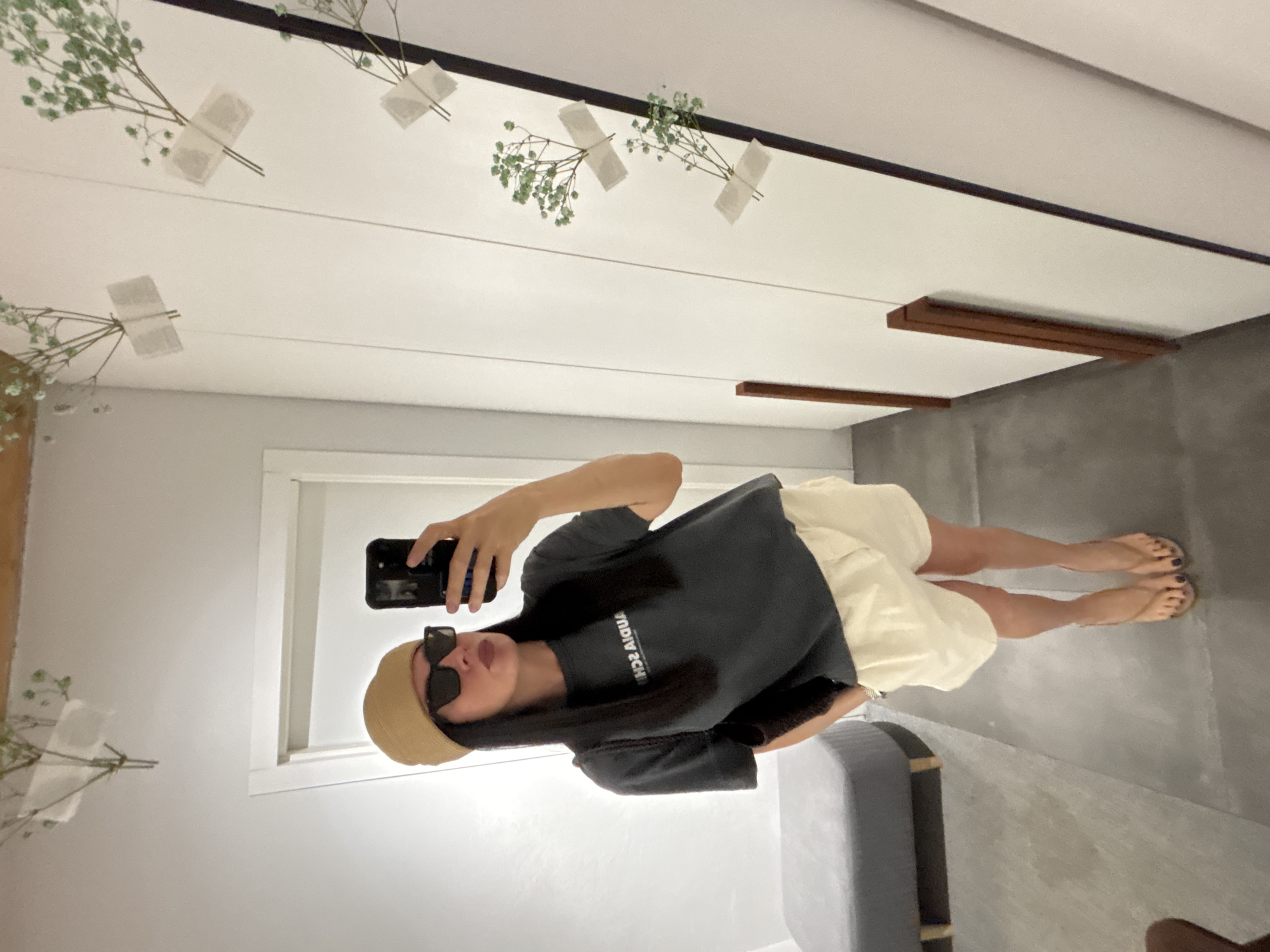 A mirror selfie depicts a young person indoors, seemingly in a store or dressing room. The individual wears a brown hat, sunglasses, a dark-colored t-shirt, and light-colored shorts. They hold a black smartphone in their right hand, taking the photograph. The setting appears to be modern and minimalistic. The person's reflection shows part of their legs and feet, wearing flip-flops or sandals. The image gives the impression of an informal, personal moment captured by the subject themselves. There is no indication of the city of Chaikovsky, Russia.