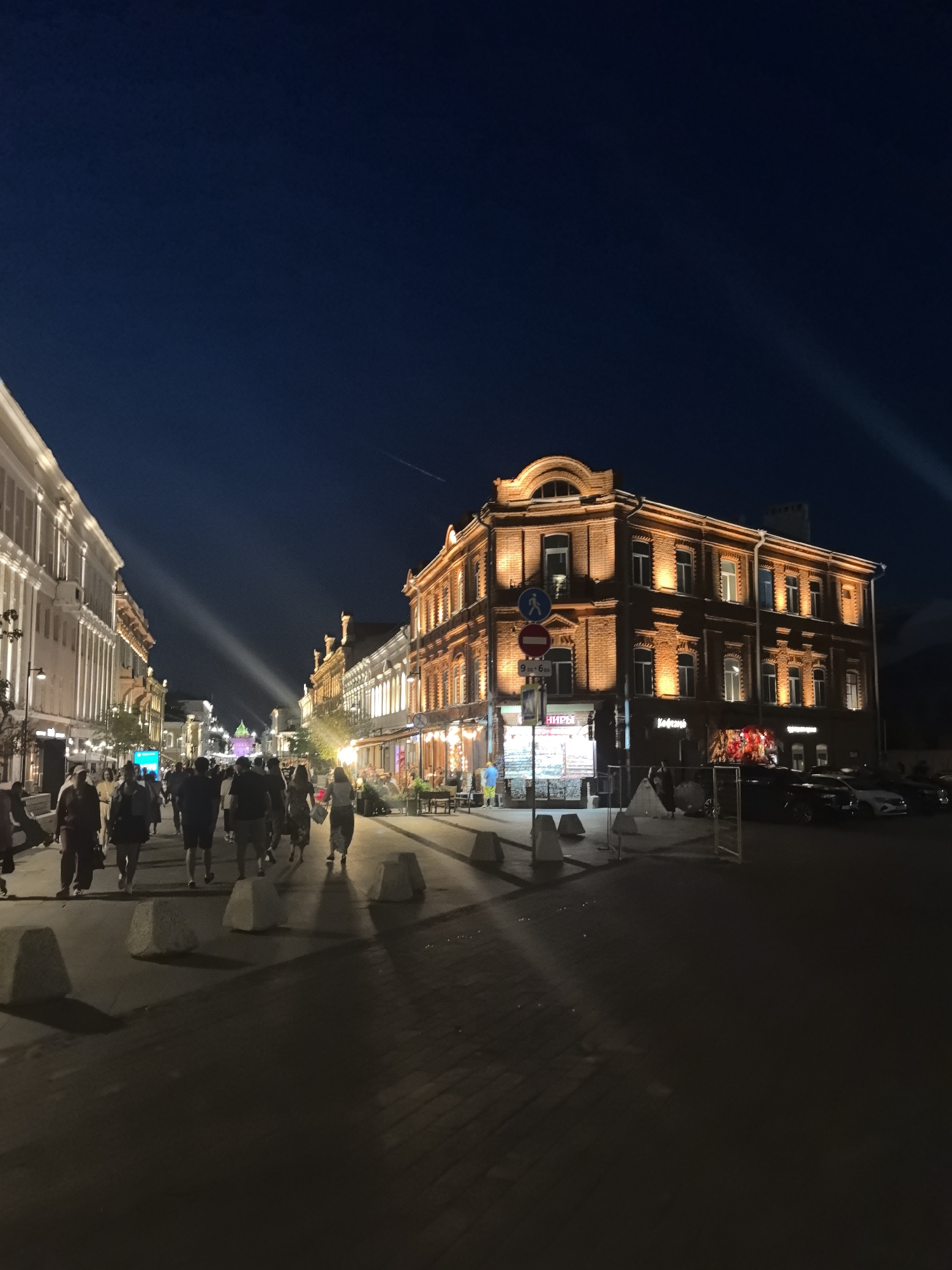 The image depicts a street scene at night in Nizhny Novgorod, Russia, likely in the historical center. The foreground shows a paved pedestrian walkway with several concrete bollards casting shadows. A group of people are walking away from the camera, their forms silhouetted against the ambient light.
To the right, a multi-story brick building is brightly illuminated, highlighting its architectural details, including windows and decorative elements. Signs are visible on the building's facade, one appearing to read "Кофейня" (Coffee shop) and another possibly indicating business hours. Cars are parked along the street in front of the building.
The left side of the image shows another building with a lighter colored facade, featuring vertical architectural lines. Further down the street, more buildings line both sides, with some displaying illuminated signs and window displays. A bright, narrow beam of light cuts across the scene from the upper left, suggesting a spotlight or projector. The sky above is a deep blue, indicative of night. The overall impression is that of a lively, well-lit urban street at dusk or early evening.