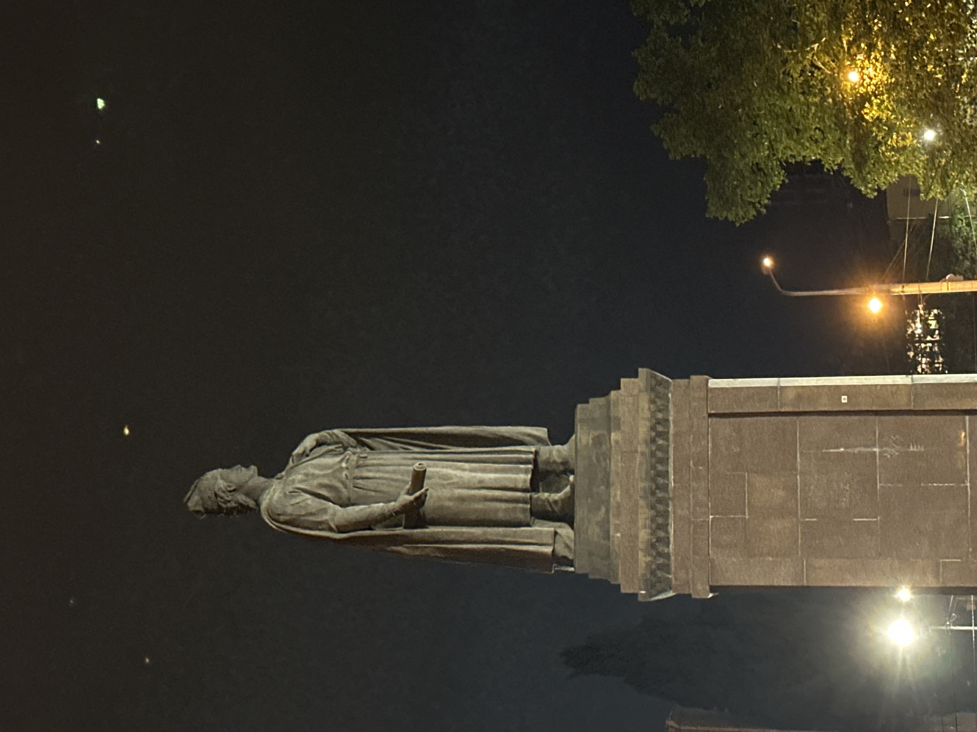 Here is a detailed description of the image:
The image shows a statue at night. The statue depicts a man in a robe, holding a scroll or cylinder in his hand, standing on a large rectangular stone pedestal. The pedestal has several tiers and is made of stone blocks. In the background, there are dark trees and streetlights, suggesting an urban park or city square. The sky is dark, indicating that it is nighttime. The statue is likely located in the city of Unknown, Unknown, as specified in the prompt. The statue and its pedestal are the main subjects, illuminated by streetlights. There are no people visible other than the representation in the statue. The environment appears to be a public space where the statue is erected.