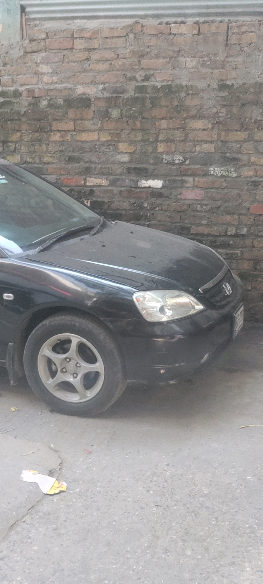 A black car is parked in front of a brick wall with corrugated metal sheeting above it. The car's hood and front wheel are visible. The car is dusty, and there is some debris on the ground in front of it. The scene appears to be an outdoor urban setting, possibly in the Metropolitan Corporation Rawalpindi, Pakistan, given the context. The brick wall is old and weathered, with some green moss growing on it. The ground is paved with asphalt. There is a license plate on the front of the car, but the text is illegible.