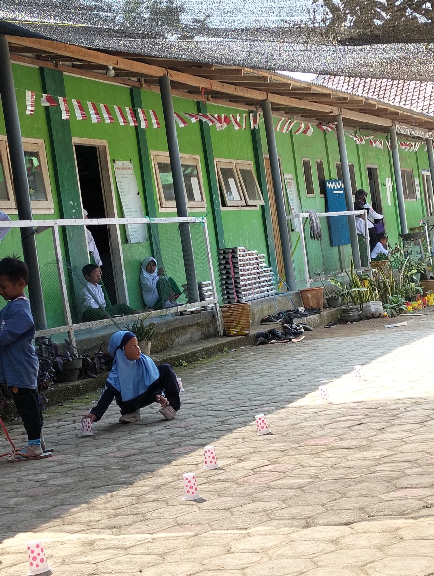 In Desa Wringin Putih, Indonesia, a group of children are engaged in an outdoor activity in front of a green, single-story building. The building has a wooden overhang supported by posts and is decorated with red and white pennant banners. Several windows are visible along the building's facade, some open, revealing glimpses of the interior. In the foreground, a young boy in a blue shirt and black pants stands on wooden sandals, holding a red string. Beside him, a girl in a light blue hijab and dark pants is crouching, reaching towards several small, white cups with pink polka dots scattered on the paved ground. Other similar cups are also visible further away on the ground. In the background, within a fenced area, two children are seated, one in a light blue hijab and the other in a light blue shirt. Various items are placed along the building's exterior, including stacks of what appear to be trays, baskets, and potted plants. Several pairs of shoes are also visible on the ground. The scene suggests an educational or recreational setting, possibly a schoolyard, bathed in sunlight.