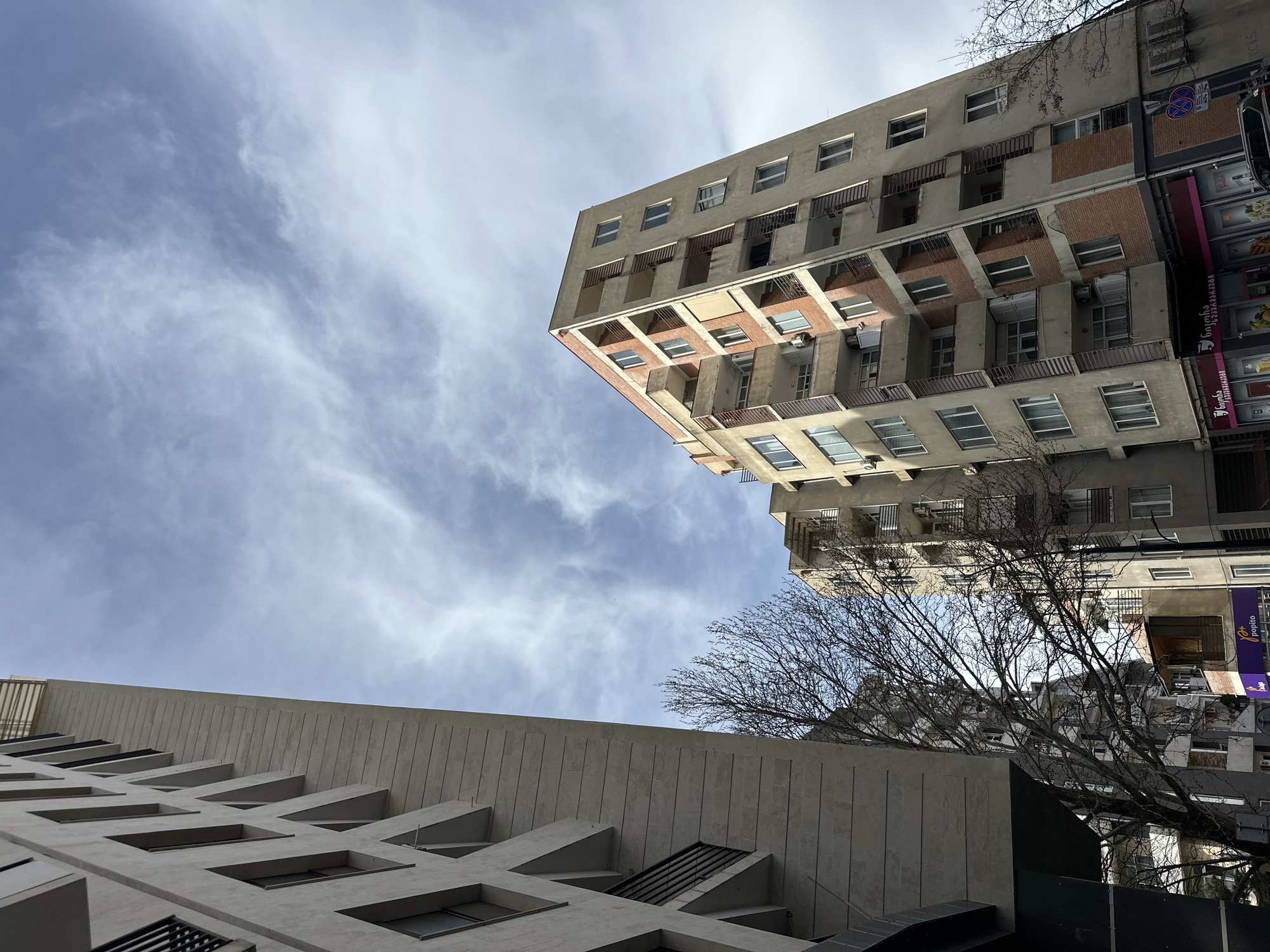 The image, taken from a low angle in Tbilisi, Georgia, captures two buildings against a cloudy sky. The building on the right is a multi-story concrete structure with repetitive window patterns and balconies. The lower building is also concrete, featuring angled windows. A bare tree is situated between the buildings. Signage is visible on the right, indicating commercial establishments. The sky is predominantly blue with thin, wispy clouds. The scene is urban.