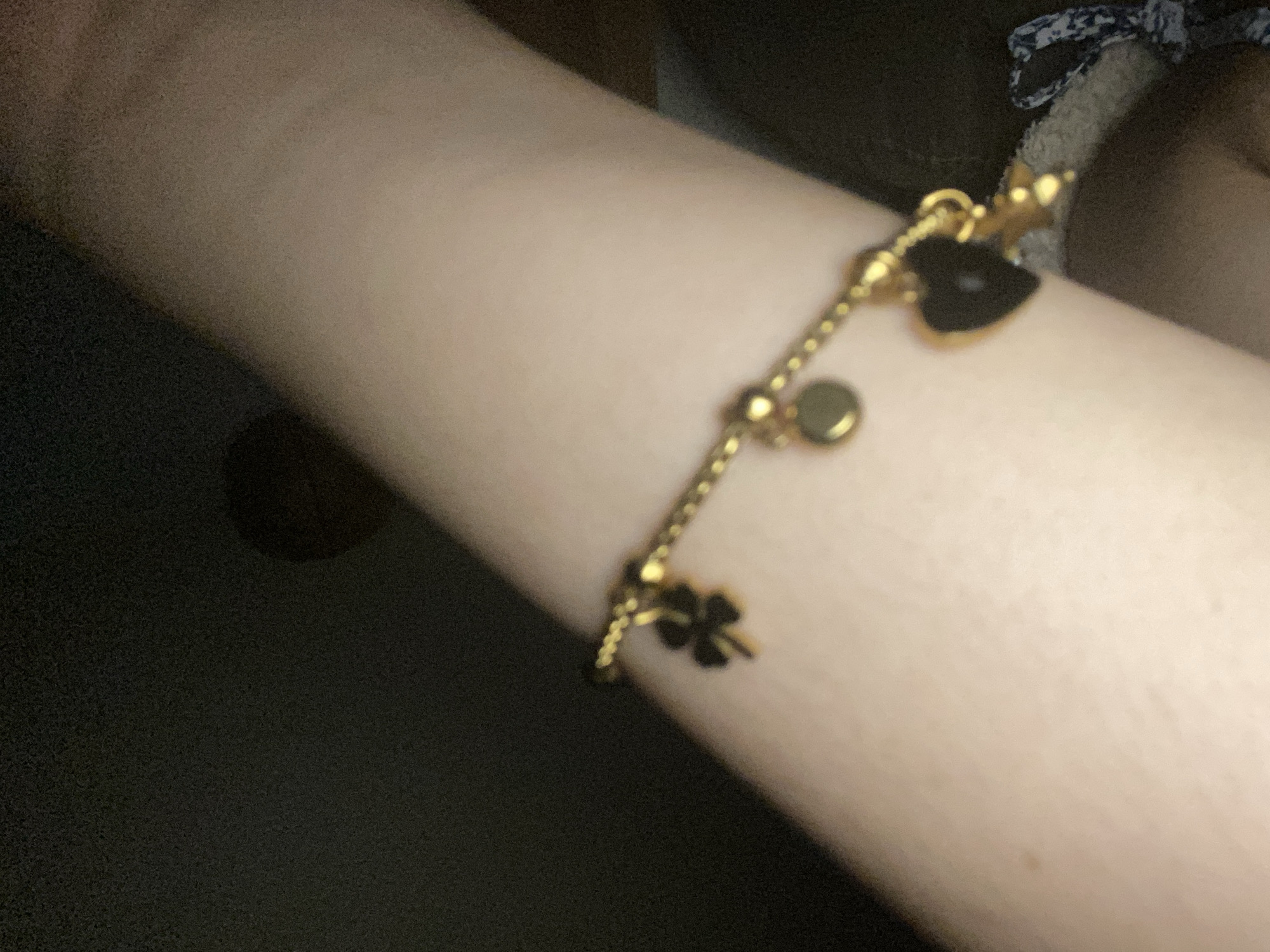 The image presents a close-up, softly lit shot of a person's inner forearm or wrist, showcasing a delicate, gold-colored chain bracelet. The arm has fair skin and is positioned horizontally across the frame.
The central subject is the bracelet, which is adorned with several charms. Visible charms include a distinct dark, heart-shaped pendant, a small circular gold bead, and a dark, possibly four-leaf clover shape. The chain itself appears to be made of small, interconnected beads or links.
The background is predominantly dark and out of focus, indicating an indoor setting with low ambient light. A small, blurry portion of patterned fabric, featuring a dark base with lighter, abstract shapes, can be glimpsed in the upper right corner, likely part of clothing worn by the person.
The activity depicted is simply the wearing and display of the bracelet. Due to the dim illumination, the scene suggests it might be evening or night. There are no specific location cues visible within the image itself to indicate Cotoperíz, Venezuela, nor are any emotions discernible. No text is visible in the media file.