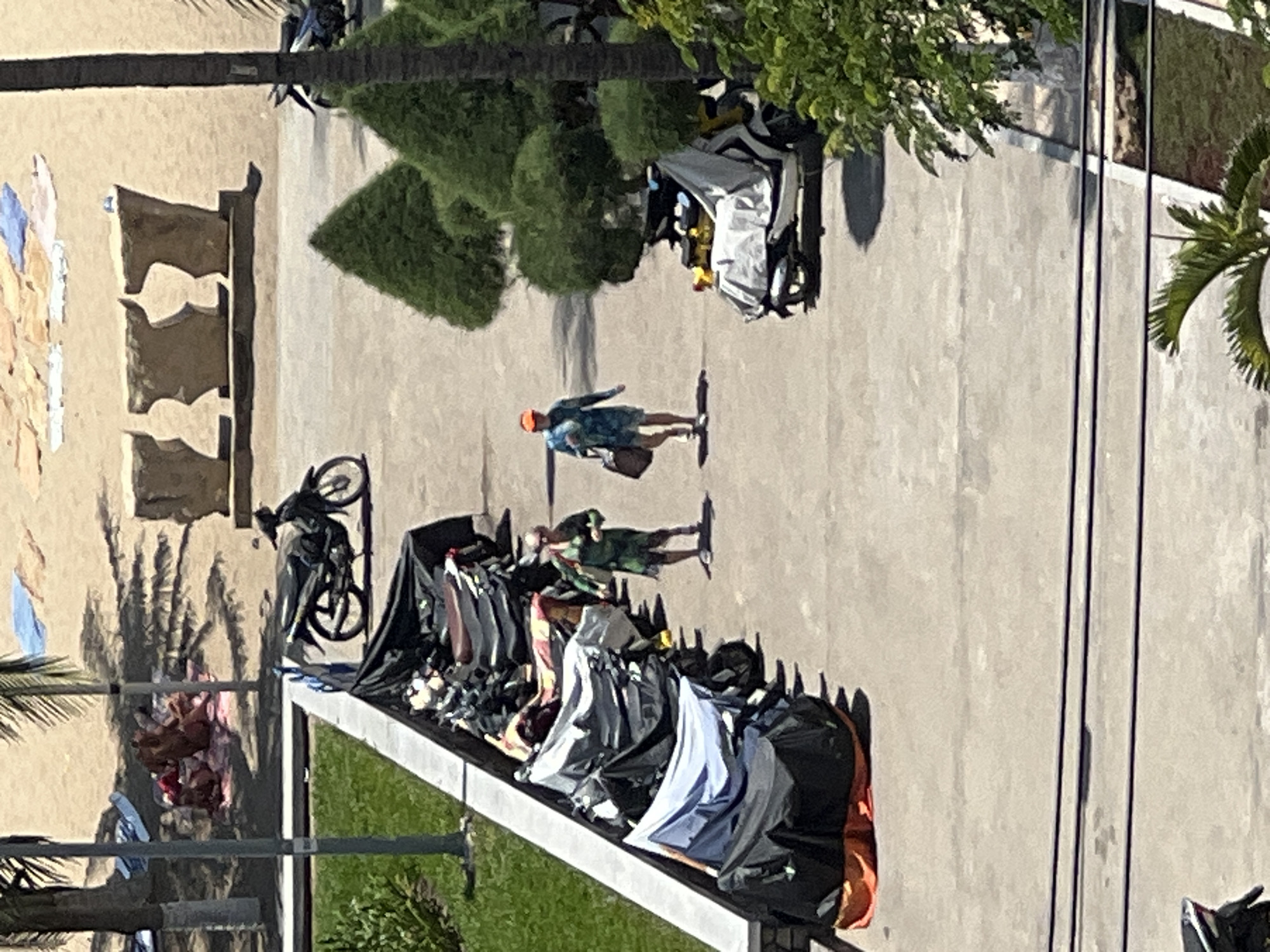 The image depicts a street scene in Nha Trang, Vietnam, near a beach. In the foreground, a row of motorcycles, mostly covered with tarps, are parked along a paved area. Two individuals are walking in the middle ground, one wearing a green outfit and the other a blue patterned shirt and shorts with an orange hat, each carrying a bag. In the background, the sandy beach is visible with scattered beach towels and a group of people lounging. Palm trees and manicured bushes are present throughout the scene, suggesting a tropical or subtropical coastal environment. Overhead power lines are also visible in the lower portion of the image. The lighting indicates a sunny day.