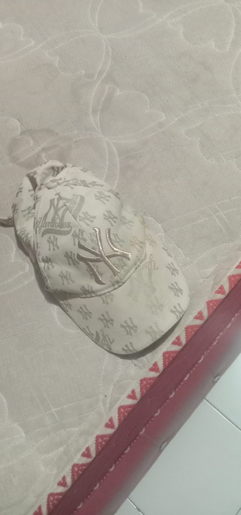 A white New York Yankees baseball cap is positioned on a white textured surface, possibly a mattress or a quilt, with a red patterned border visible on the right side. The cap has a repeating pattern of the "NY" logo and the word "Yankees" in a light gray color. The main "NY" logo on the front of the cap is a slightly darker shade of gray and appears to be embroidered. The surface the cap rests on has a floral or swirling pattern stitched into it. A red border with a triangular pattern in white and red is located along the right edge of the image. The background is out of focus but appears to be a light-colored tiled floor. This scene is likely in a residential setting within the city of Kendari, Indonesia.