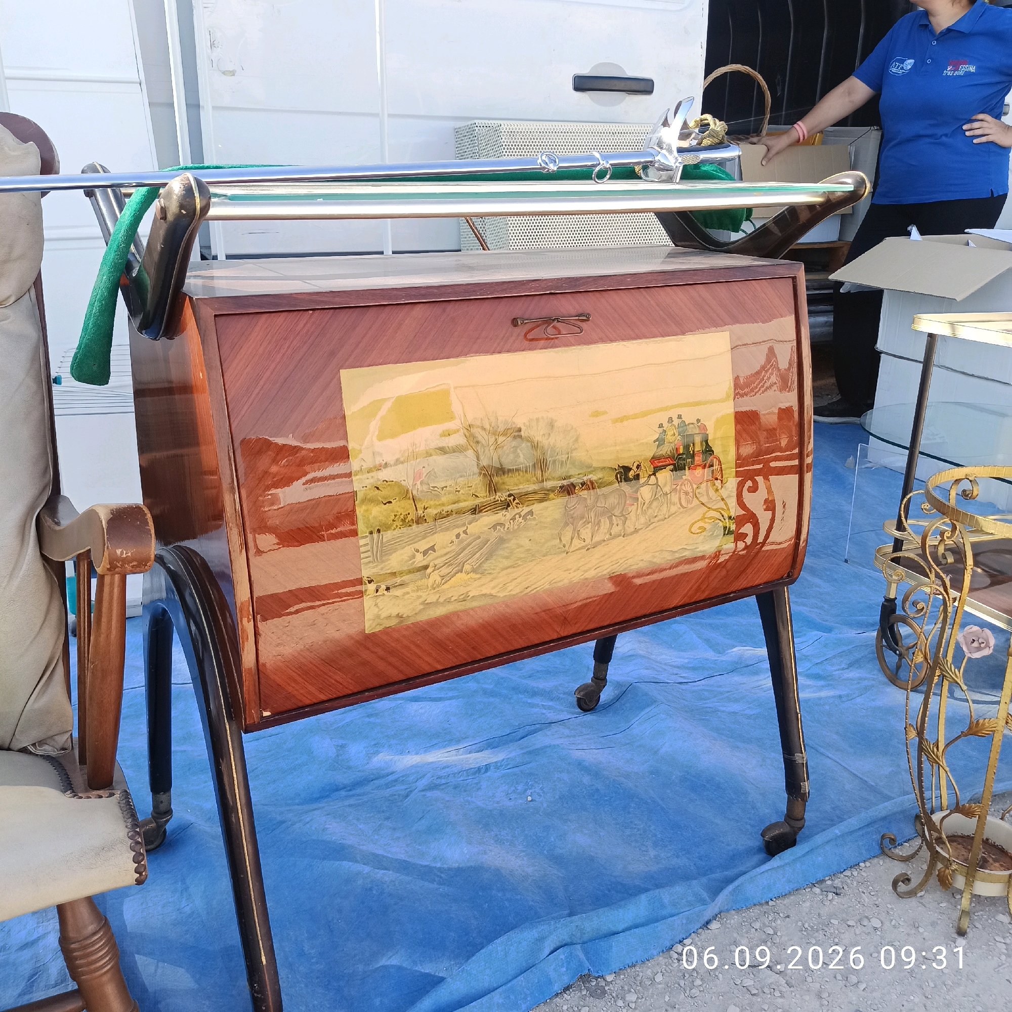 In Sannicandro di Bari, Italy, this image captures a close-up of vintage furniture items placed outdoors on a blue tarp. The primary subject is a mid-century style wooden cabinet with a glossy, reddish-brown finish, featuring a decorative panel on its front depicting a horse-drawn carriage scene with figures and dogs in a pastoral setting. The cabinet is supported by dark, slender legs with casters. To the left, partially visible, is a wooden armchair with a light-colored upholstered seat and back, adorned with decorative nailhead trim. A green fabric item hangs over the arm of the chair and is draped over a chrome rail that extends above the cabinet. In the background, a white van is visible, suggesting a potential sale or relocation of these items. To the right, a decorative gold metal stand with glass shelves and floral motifs, possibly a plant stand or etagere, is also present. A person wearing a blue polo shirt and dark pants stands in the background to the right, near the van, with their right hand on their hip. A woven basket is visible behind the person. The ground is covered with a blue tarp, with scattered gravel and debris visible at the bottom right. A timestamp "06.09.2026 09:31" is superimposed on the lower right portion of the image.