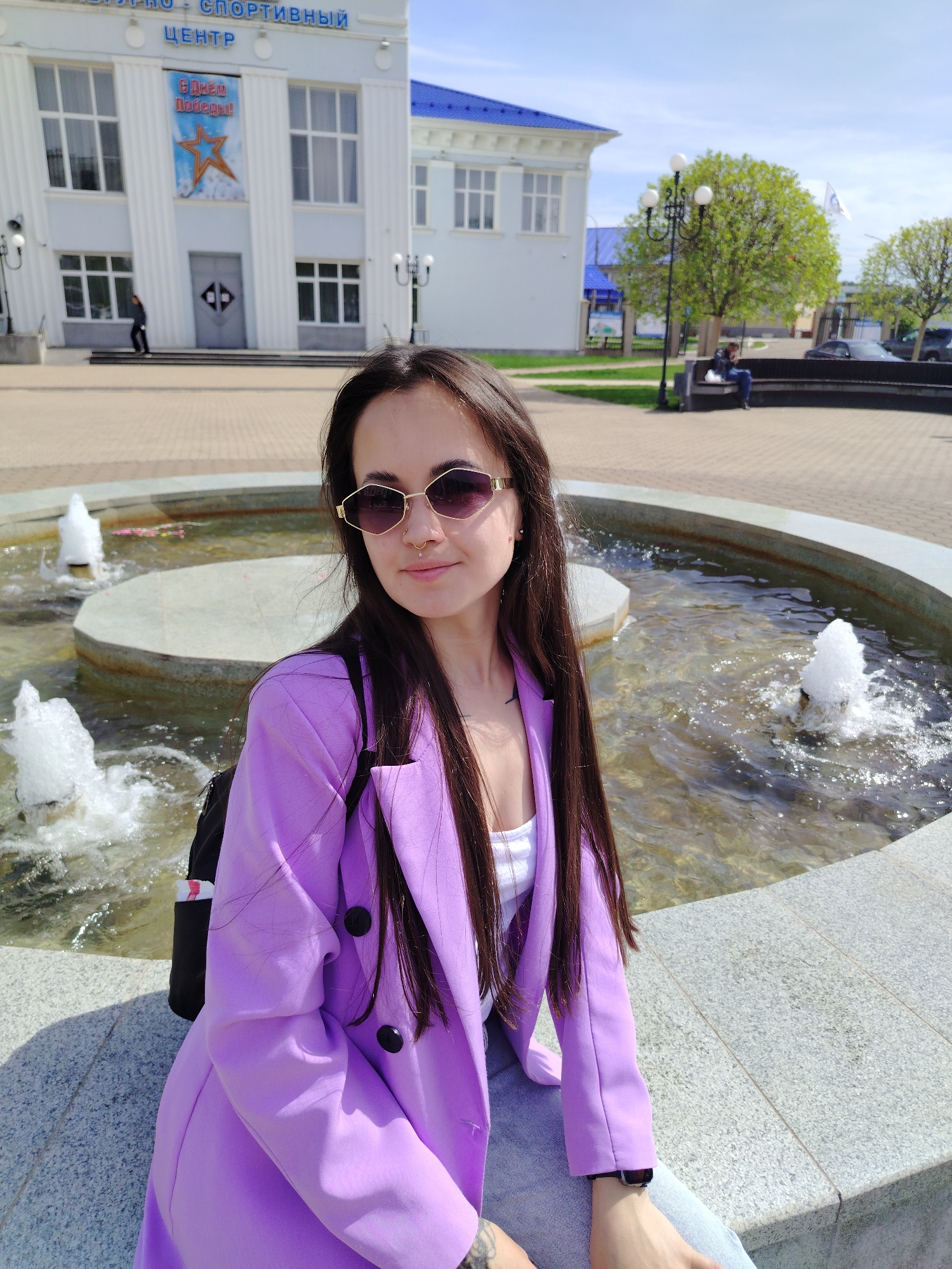 A young woman with long, straight dark hair and sunglasses sits on the edge of a round fountain in the city of Chaikovsky, Russia. She is wearing a lilac blazer and a white top. Behind her is a water fountain with multiple spouts and a building. The building is white with a blue roof and features the inscription "Центр" and a red star. Trees, street lamps, and other buildings are also visible in the background. The ground is paved with light brown bricks.