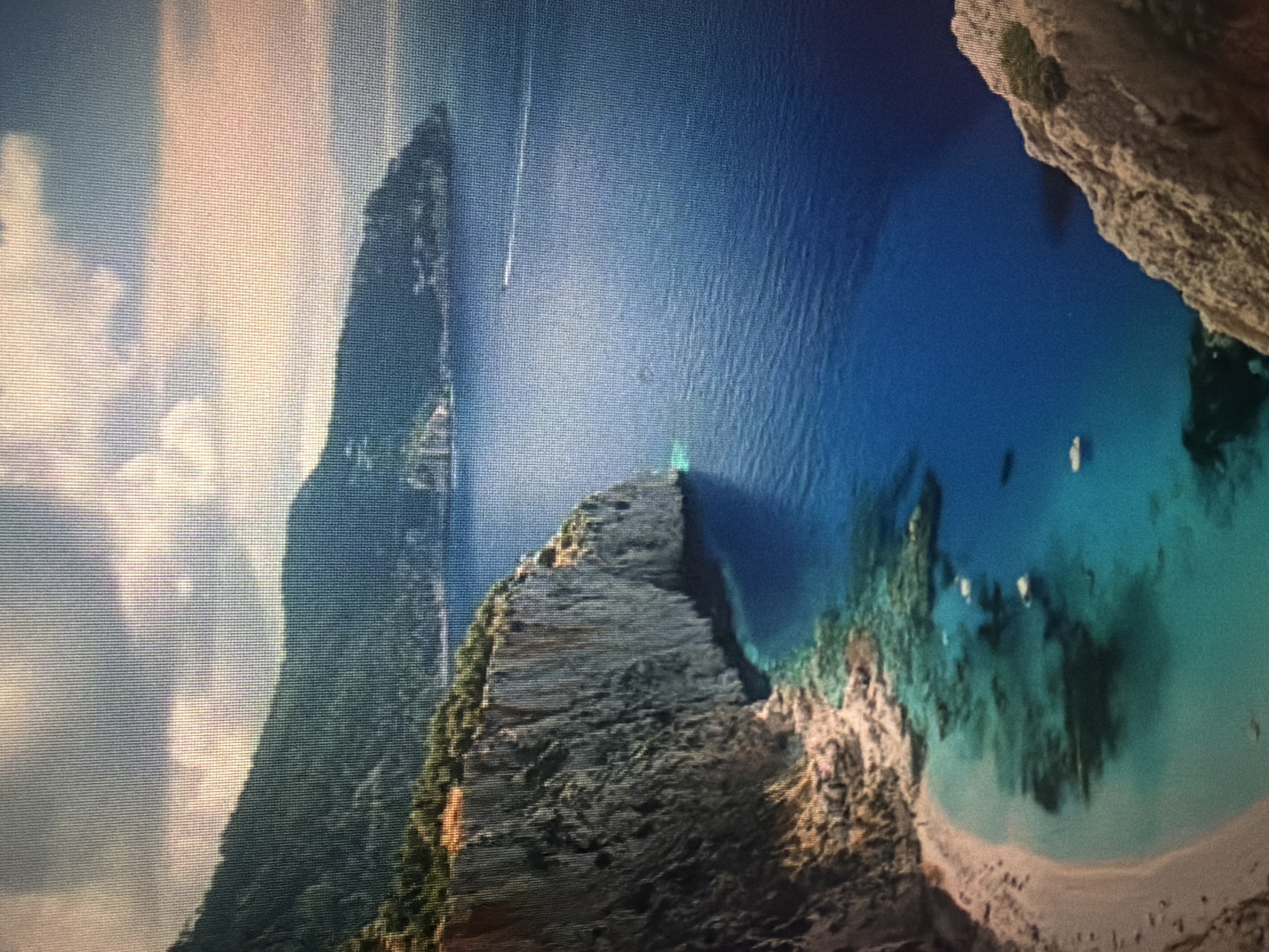 The image displays a picturesque coastal landscape featuring dramatic cliffs overlooking a bay with turquoise waters. Lush green vegetation covers the tops of the cliffs and the distant mountains. The sea is a deep blue, with a small white boat visible in the distance. A sandy beach curves along the edge of the bay, with clear water revealing underwater rocks and shadows. The scene is bathed in sunlight, with some clouds present in the sky. There is no indication of the city of Baan Mae Nam, Thailand in the image.