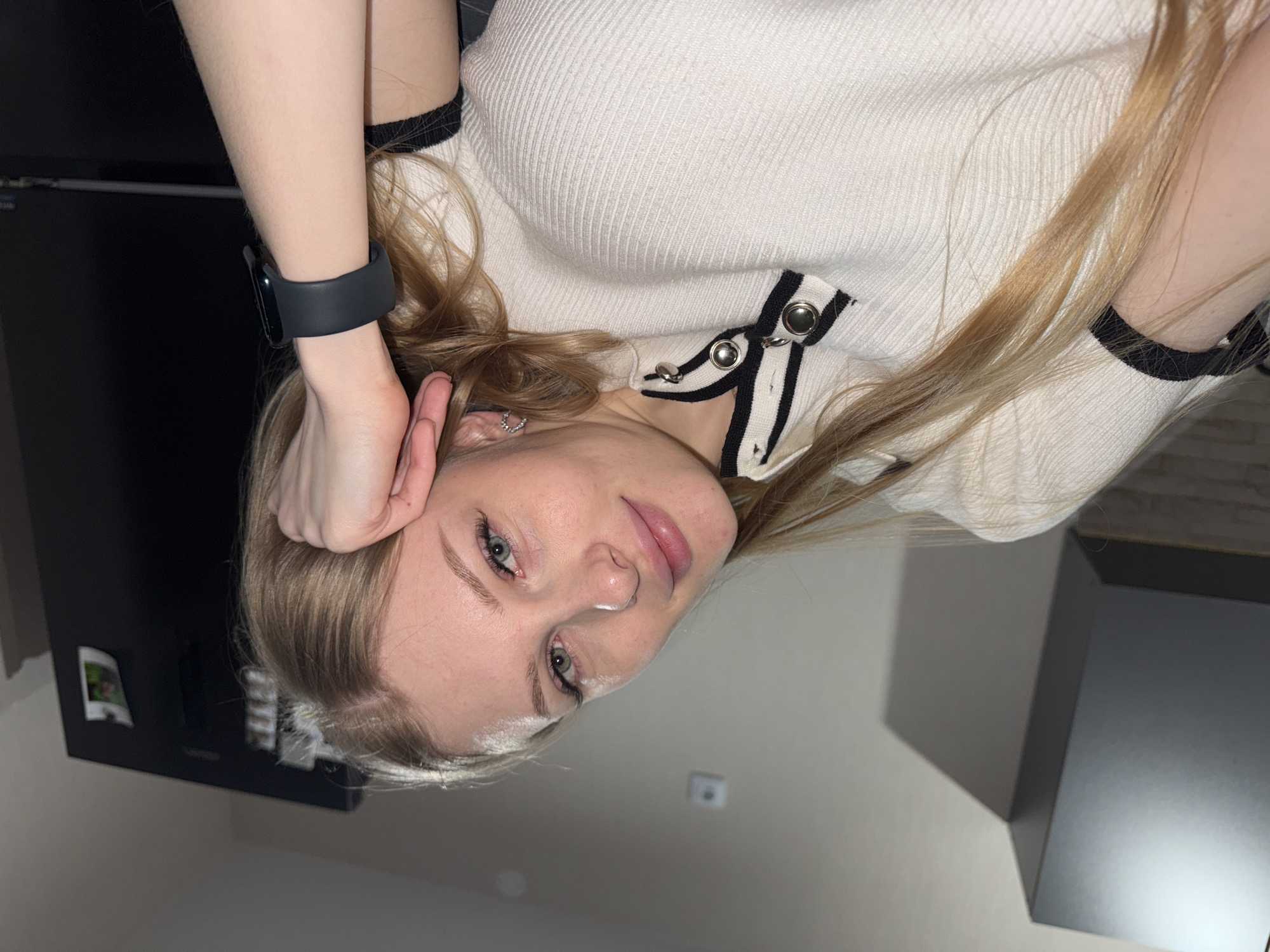 A young woman is posing for a selfie. She is wearing a white ribbed top with black and white accents and a black smart watch. Her blonde hair is partially obscuring her face. The background includes a dark cabinet, a light-colored wall, and other indeterminate features. There is no visible evidence to indicate the location is in the city of Perm, Russia.