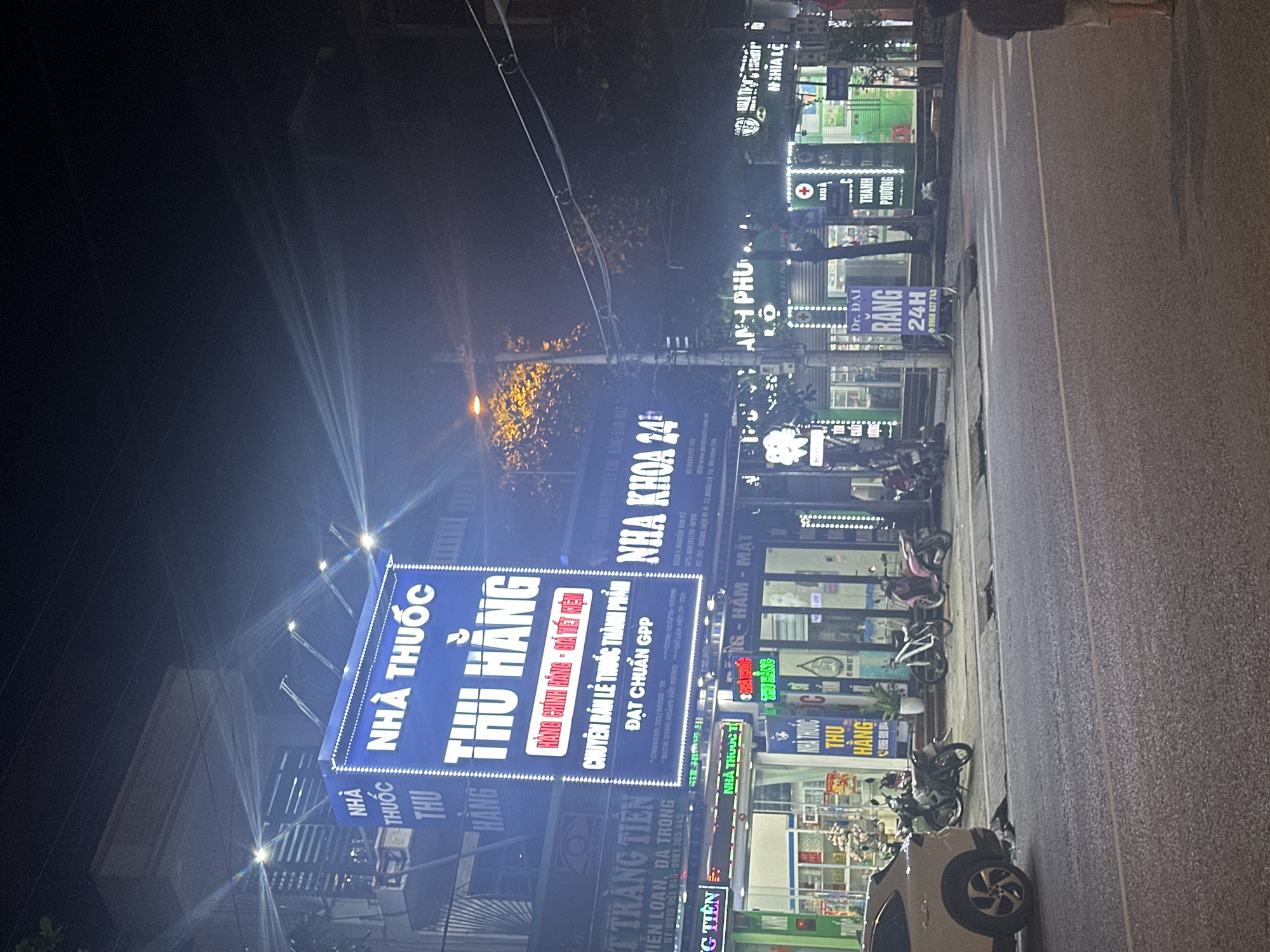 This night-time image depicts a commercial street in Trung Tam Ward, Vietnam. The scene is illuminated by numerous bright neon signs and streetlights, generating prominent lens flares across the upper portion of the photograph.
In the foreground, an asphalt road with a faint white center line is visible. A concrete sidewalk lines the right side of the road, where several motorbikes and one bicycle are parked. A white SUV is partially visible on the far left. On the right, a person in dark clothing and light shorts stands with their back to the viewer, at the edge of the road.
The midground is dominated by multiple brightly lit storefronts. A large, rectangular blue sign for "NHÀ THUỐC THU HẰNG" (Thu Hang Pharmacy) is prominent on the left, displaying white Vietnamese text indicating "Genuine Goods - Economical Prices" and "Specializing in Retail of Finished Medicines, GPP Standard Compliant." Below this, another smaller "NHÀ THUỐC THU HẰNG" sign is visible. To its right, a sign for "NHA KHOA 24H" (24H Dentistry) is displayed. Further right, a pharmacy with a green facade and a red cross symbol, labeled "THANH PHƯƠNG," is visible. A smaller, vertical purple sign on the sidewalk reads "Dr. ĐẠI RANG 24H" (Dr. Dai Dentistry 24H). Other businesses, including "TRANG TIỀN," are partially visible. Overhead, utility poles and power lines crisscross the frame.
The background consists of dimly lit building structures and trees, silhouetted against the dark night sky. The overall impression is that of a bustling urban commercial area after dark.