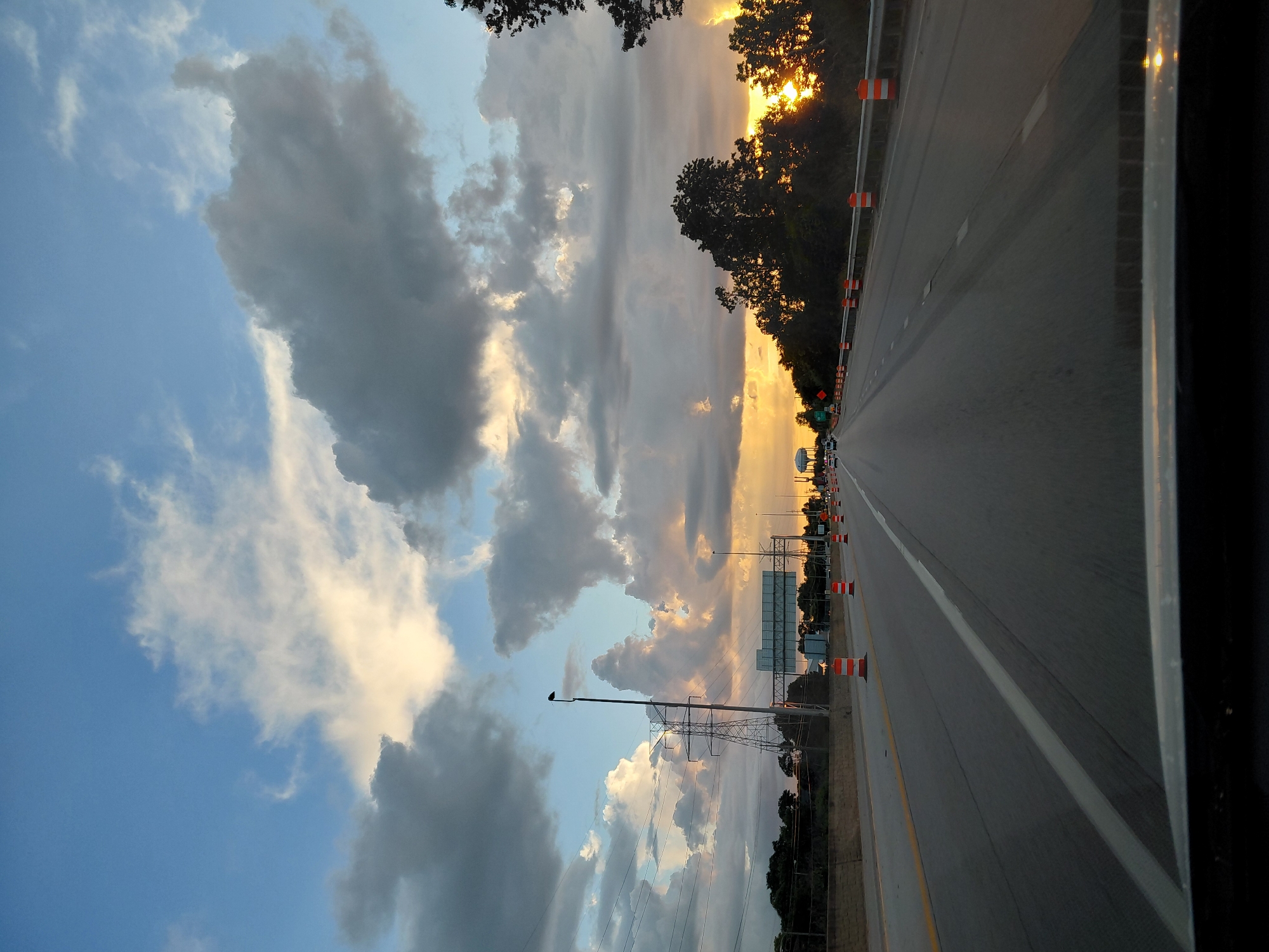 This is a photograph of a highway in Lexington, Kentucky, USA, taken from a vehicle's perspective at sunset. The sky is filled with dramatic clouds, with patches of blue sky visible. The sun is low on the horizon, casting a warm, golden light through the clouds. On the right side of the highway, there are trees silhouetted against the sunset. The highway itself has multiple lanes, with traffic cones indicating construction or a lane closure. A tall electrical tower and power lines are visible on the left side of the highway. The road surface is asphalt, with white lane markings. The image appears to be a snapshot of a typical evening commute on a major road.