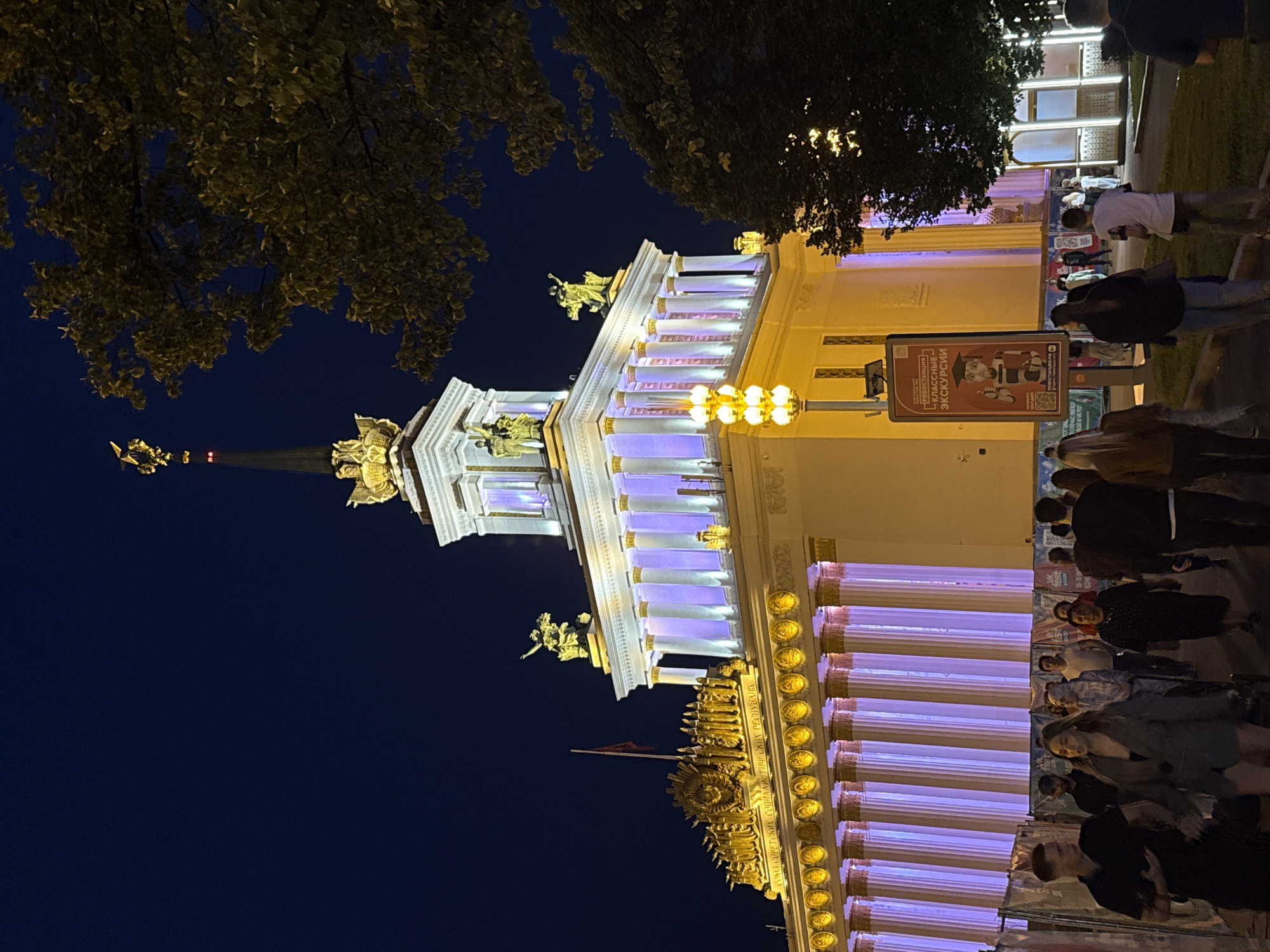 This nighttime image captures the monumental VDNKh (Exhibition of Achievements of National Economy) complex in Moscow, Russia. The central structure, an exhibition pavilion, is dramatically illuminated with purple lights accentuating its classical architecture, complete with rows of columns topped by golden sculptures and a towering spire adorned with a golden figure. Several people are walking along a paved area in front of the pavilion, some illuminated by streetlights and the building's glow. A large poster featuring a robot with text in Russian is prominently displayed on a pole to the right of the pathway. Trees with dense foliage are visible on the right side of the frame, partially obscuring the pavilion and contributing to the atmospheric nighttime setting. The sky is a deep, dark blue, indicating it is late evening or night.