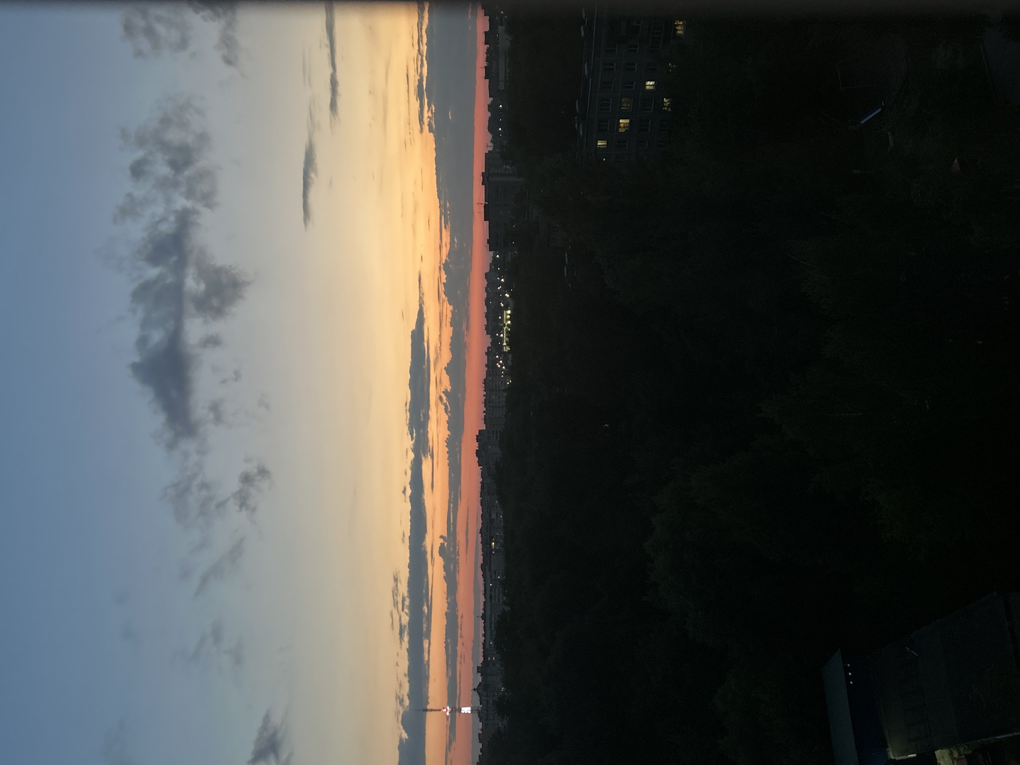 An aerial shot captures a cityscape at dusk. The sky transitions from a deep blue at the top to shades of orange and pink near the horizon, where scattered clouds drift. A tall communication tower with a lit top stands prominently in the distance, surrounded by buildings of varying heights. Dark, dense trees dominate the middle ground, obscuring much of the city below. In the foreground, the dark silhouettes of two low-rise structures are visible, one with a visible roofline. The overall impression is of a quiet urban twilight.