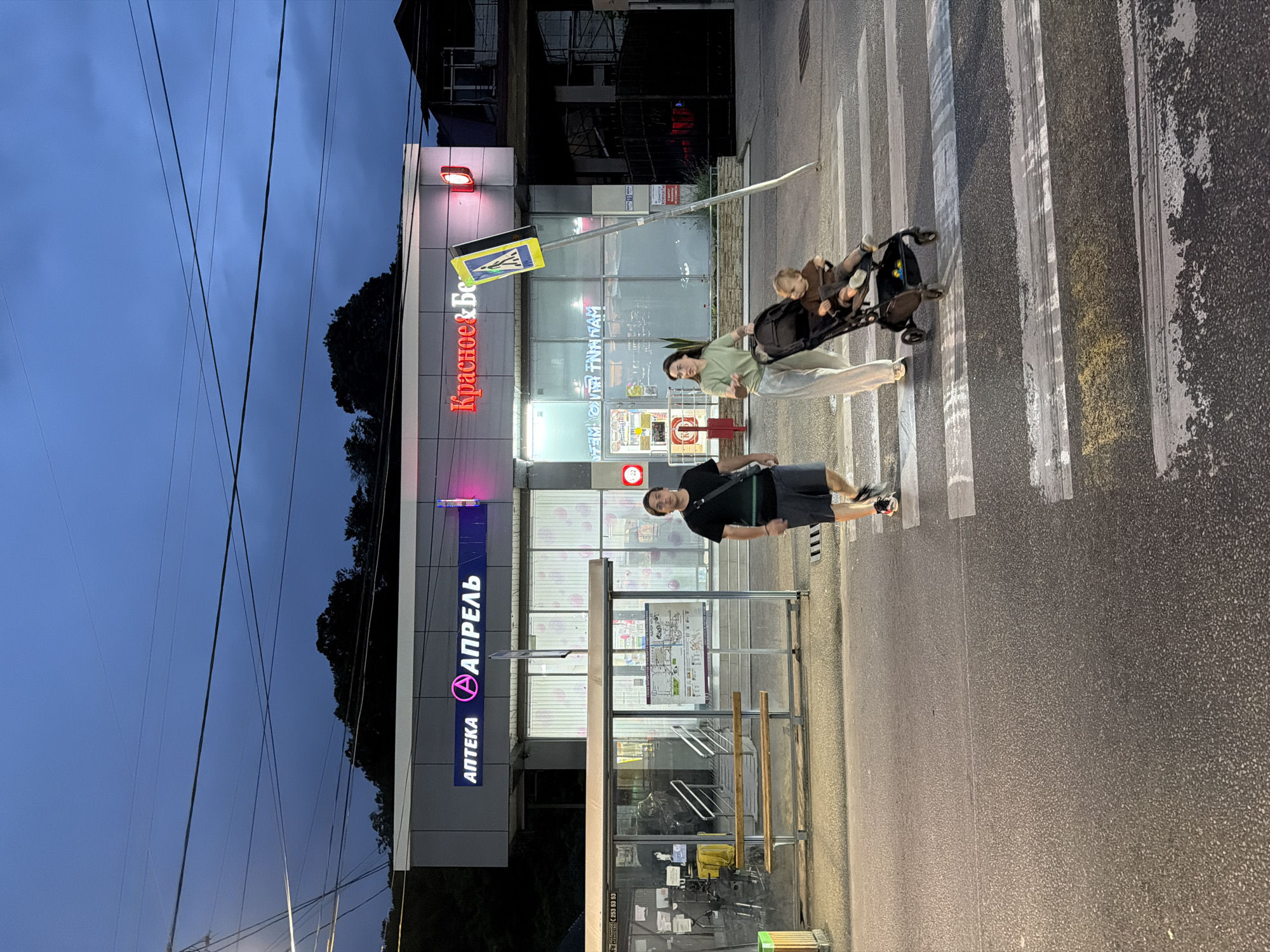 A family crosses a pedestrian street at dusk in Sochi, Russia. A man, woman, and child are captured mid-stride. The man, wearing a dark t-shirt and shorts, walks ahead on the right side of the crosswalk. The woman, in a light green top and beige pants, pushes a stroller containing a young child who is facing forward. The stroller is dark-colored and equipped with a sunshade. They are crossing a well-lit street with visible white pedestrian markings. In the background, two storefronts are illuminated, one with Cyrillic signage reading "АПТЕКА АПРЕЛЬ" (Pharmacy April) and another with red neon lettering that appears to say "Красное & Белое" (Red & White), indicating a store. A bus stop shelter with wooden benches is visible to the left of the pharmacies. The sky is dark blue with hints of clouds, suggesting the early evening. A tall, leaning street sign with a pedestrian crossing symbol stands diagonally on the right side of the crosswalk. The overall setting is urban, with overhead power lines visible against the darkening sky.