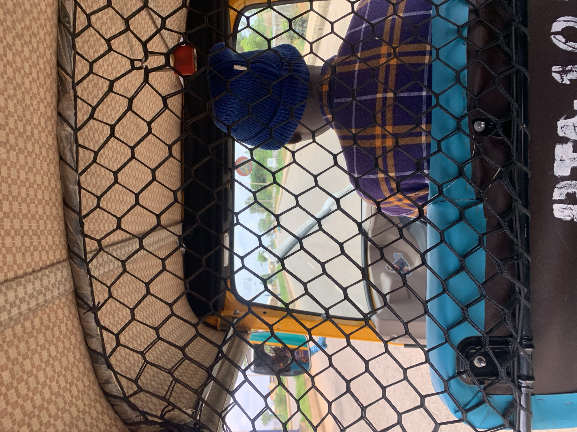 The image captures an interior view looking forward from a passenger perspective within a three-wheeled auto-rickshaw, in the city of Rafin Par, Nigeria. A black hexagonal mesh net occupies the foreground, extending from the vehicle's patterned beige checkered roof down to a turquoise-upholstered seat or compartment. Below this, a dark brown surface displays partially visible white stenciled text, "DTA-10." The vehicle's driver, seen from behind, occupies the central frame, wearing a dark blue knit beanie and a plaid shirt with a pattern of dark purple, orange, and light yellow checks. Yellow structural elements of the vehicle frame the windshield, through which a paved road is visible, stretching into the distance under an overcast sky. Green foliage and light brown earth line the road's edges. A circular road sign, red and white, is visible on the left side of the road. A side mirror on the left reflects blurred green and turquoise elements. A small red object, possibly a decorative item or light, hangs from the vehicle's interior near the roofline on the right.