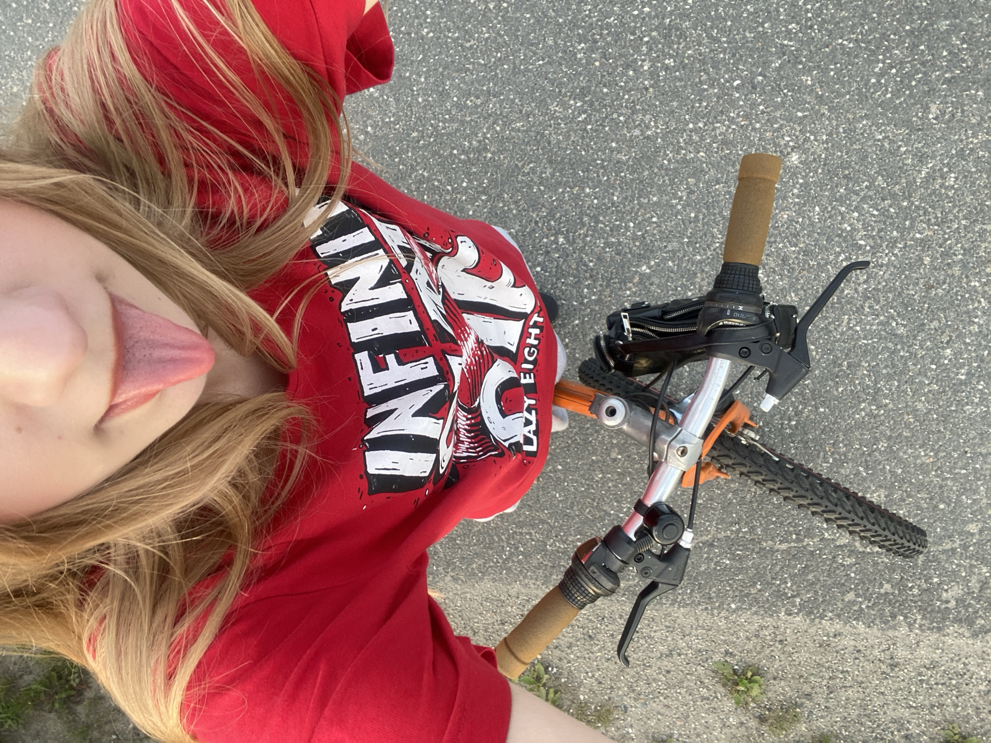 A person with long blonde hair, wearing a red t-shirt with the graphic "INFINITY LAZY EIGHT", is holding the handlebars of a bicycle. The person is sticking out their tongue. The bicycle has brown grips, a black brake lever, and a black bell. The front tire of the bicycle is visible. The ground is paved with asphalt and has some small green plants growing in it. The scene is outdoors. The location is not specified in the image or provided metadata.