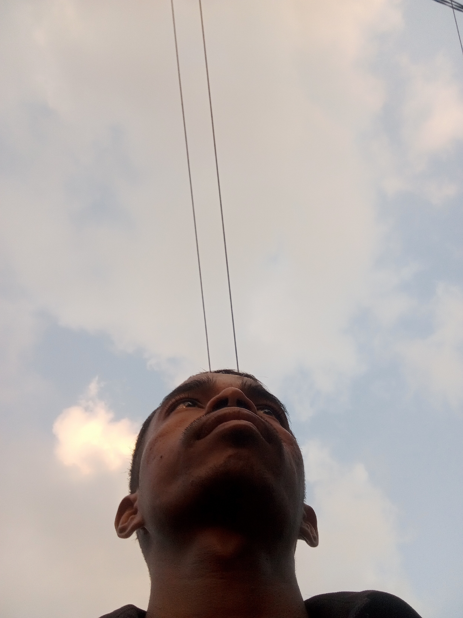 This image is a low-angle shot of a person looking upwards at the sky, with two thin wires running vertically through the frame. The person's face is partially visible, with their eyes looking towards the top of the frame. The sky is cloudy and appears to be either sunrise or sunset, with a warm glow from the sun behind the clouds on the left. The image was taken in Surabaya, Indonesia, as indicated by the location metadata. The person is wearing a dark-colored shirt. The overall impression is one of contemplation or observation of the sky.