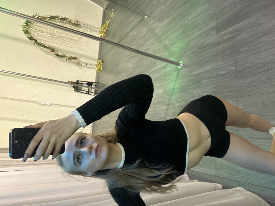 A woman with long blonde hair and blue eyes is taking a selfie in what appears to be a studio. She is wearing a black long-sleeved crop top with white trim and black shorts. A pole is visible in the background with a wreath decoration hanging on the wall. The floor is a grey tile and the lighting is bright.