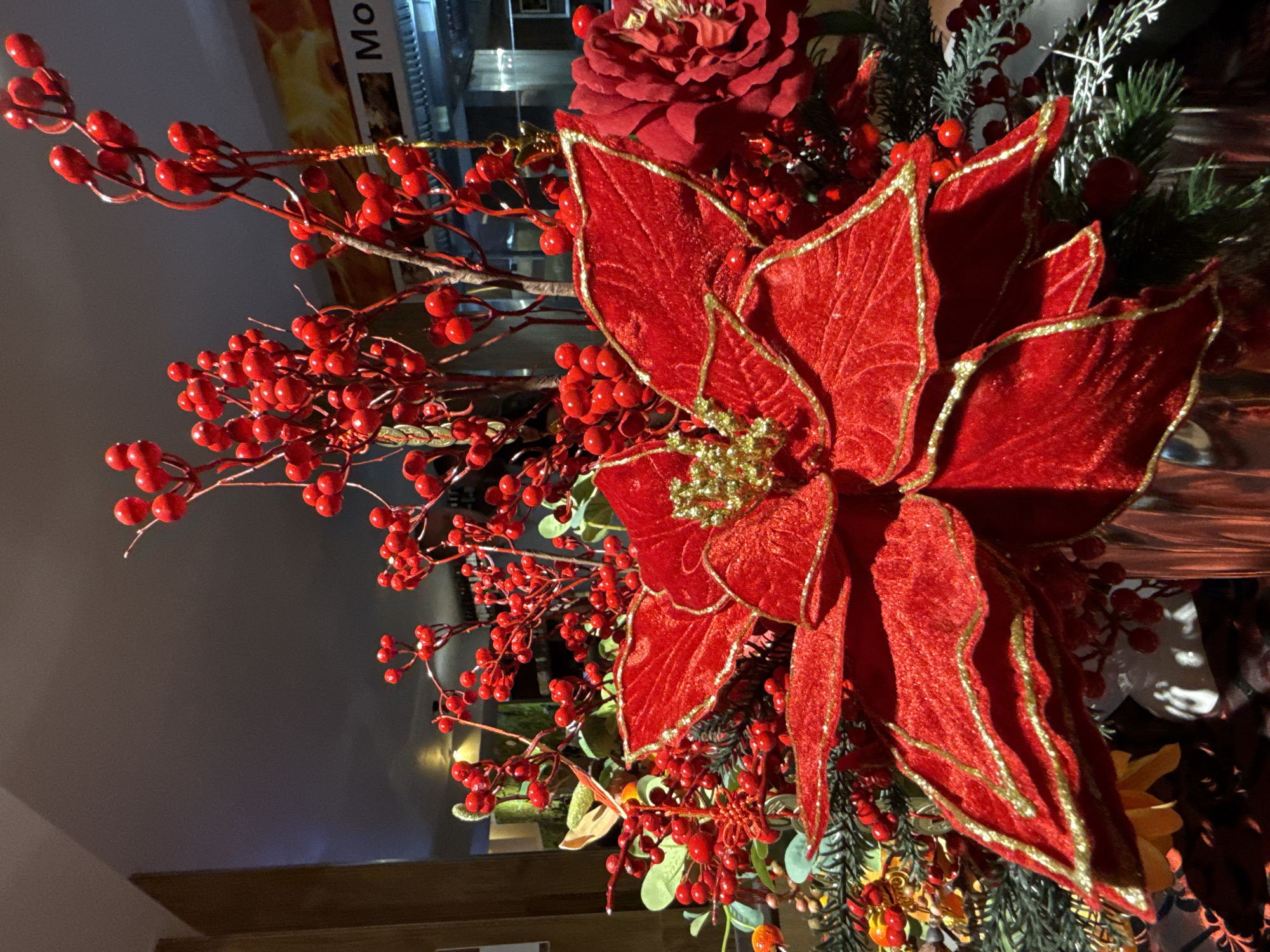 The image shows a close-up of a red poinsettia arrangement, likely a festive decoration. The poinsettia's petals are a rich red velvet with glittering gold trim, and its center is adorned with gold accents. Surrounding the poinsettia are branches with small red berries and some greenery, possibly pine or fir. In the background, a sign with the text "Mo" is partially visible, along with some out-of-focus elements of what appears to be an indoor establishment. There are no people visible in the image, and no specific location in Nordhausen, Germany, can be identified from the provided visual information. The lighting suggests an indoor setting, possibly a restaurant or a shop during the holiday season.