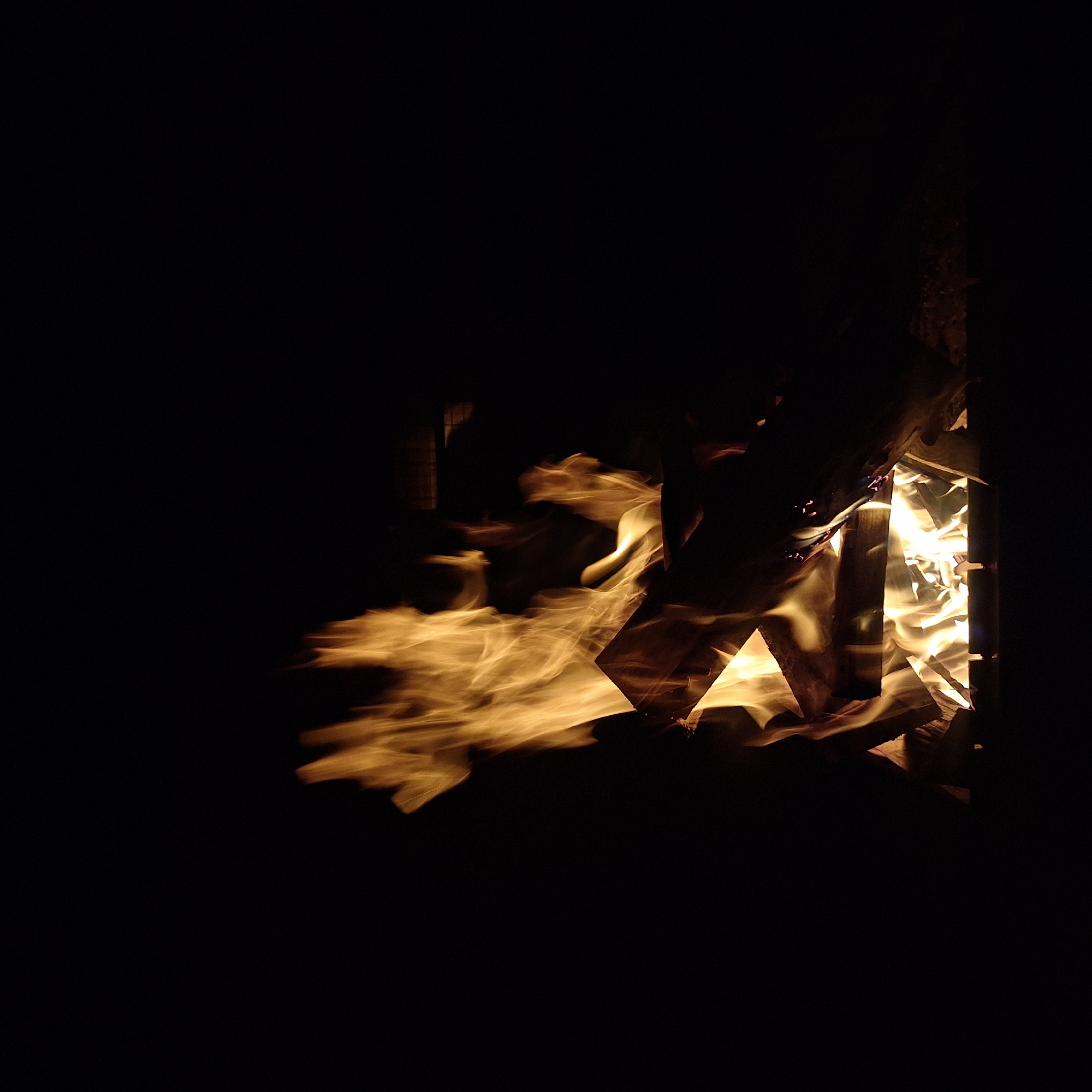 In a dark environment, likely at night, a campfire burns within what appears to be a crude enclosure. The flames are bright yellow-orange and flicker around pieces of wood. The location is Чайка Снт, Russia. No people are visible in the immediate area. The scene consists primarily of fire and wood within a dark and undefined space.