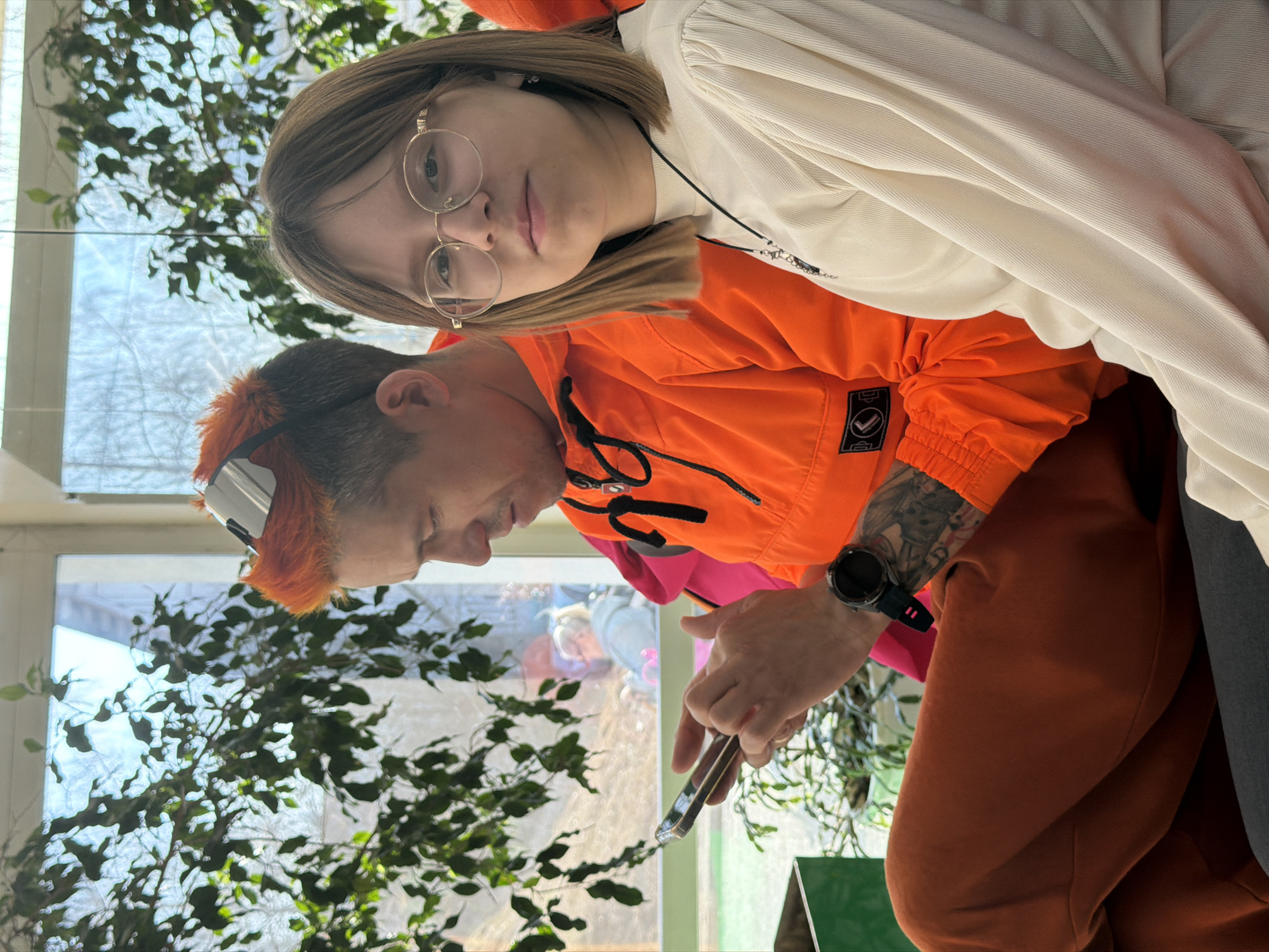 A medium, eye-level indoor shot shows two individuals in Chaikovsky, Russia. One is a person with orange hair and a matching orange jacket, holding a smartphone, likely taking a picture. This person has visible tattoos on their right arm and is wearing a black smartwatch on the same wrist. The other individual is wearing glasses and a white shirt, positioned behind and slightly above the person with the orange hair. The scene is set indoors, with a plant visible near a window. Outside the window, a vague cityscape or exterior view is discernible.