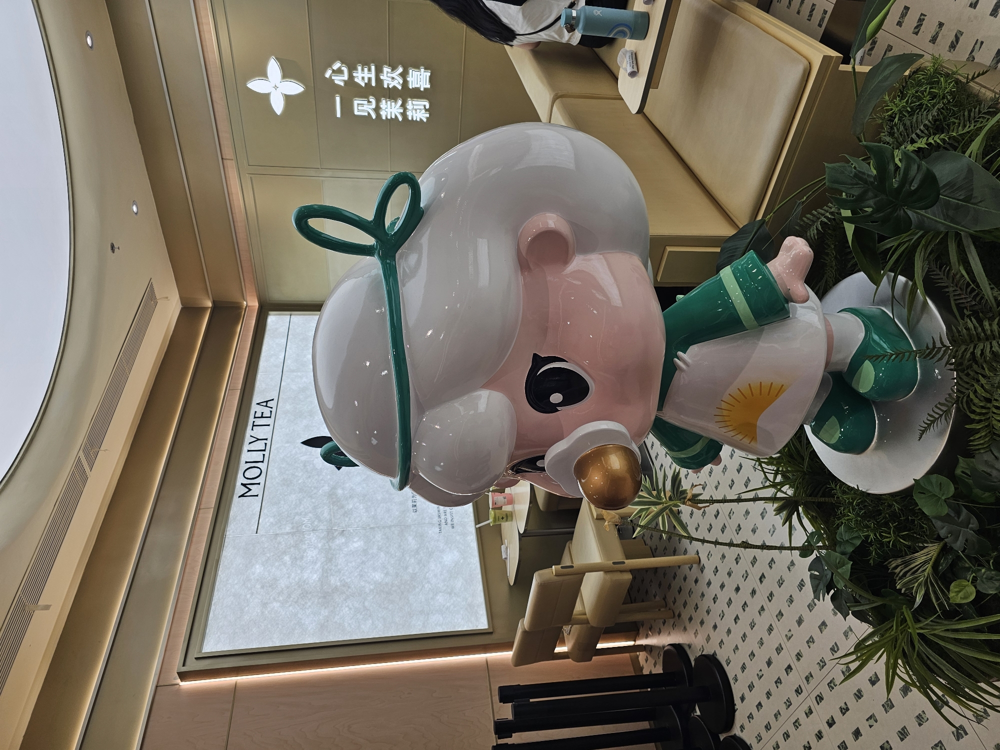 A large, cartoonish statue of a character with a white wig, green bow, and a pacifier is displayed in the foreground. The statue is dressed in a white outfit with green accents and a sun graphic on the chest. It stands on a platform surrounded by lush green plants. In the background, there is a seating area with beige chairs and tables. A wall display with the text "MOLLY TEA" is visible, along with some Chinese characters and a flower graphic. The environment appears to be a modern retail space, possibly a tea shop, with warm lighting and patterned flooring. The specific location in Condongcatur, Indonesia is not discernible from the image itself.