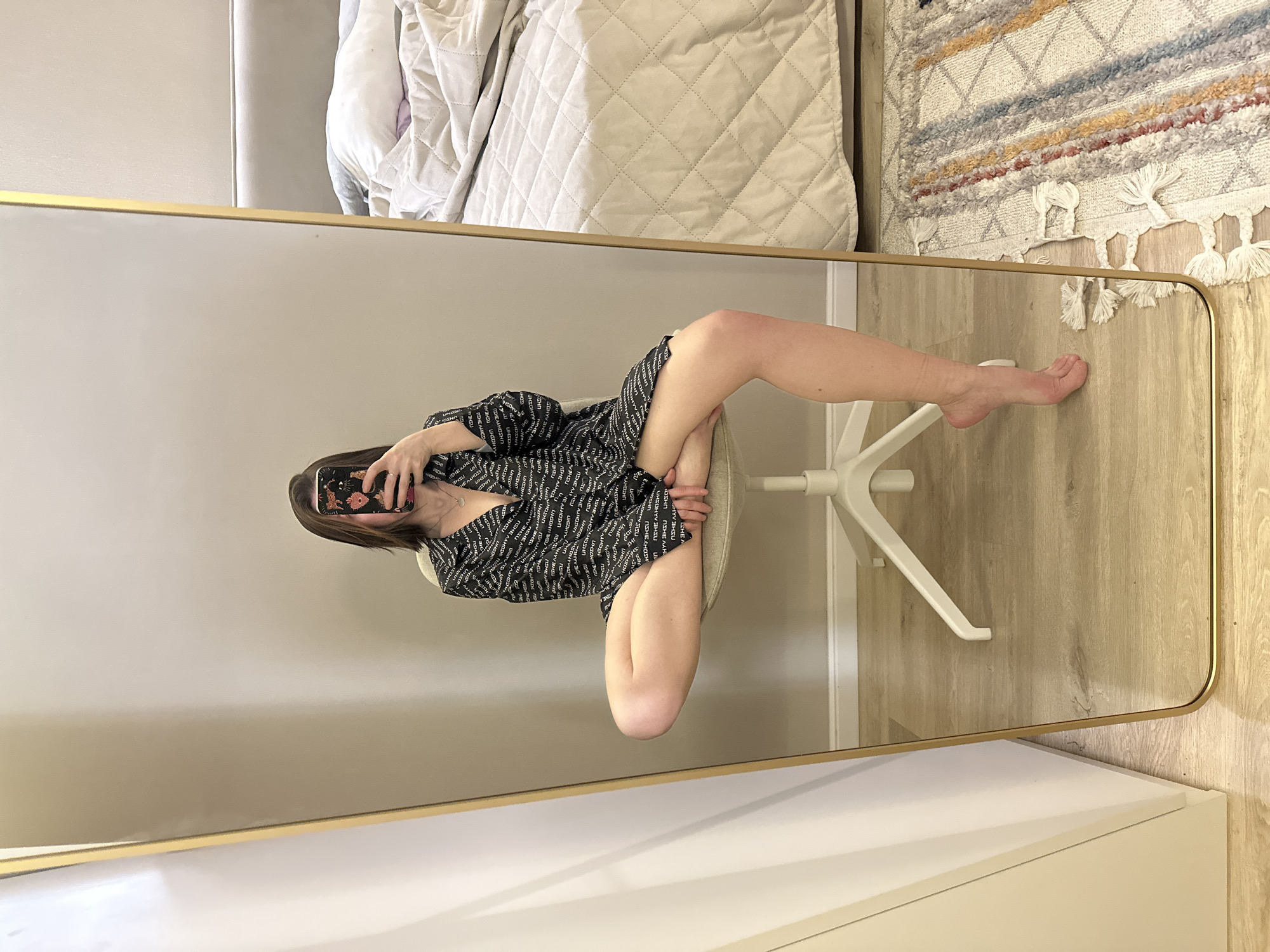 A reflection in a rectangular mirror shows a person sitting on a rolling chair. The person has a mobile phone in their hand, held at arm's length. They appear to be taking a selfie. The individual is wearing a patterned black and white top. One leg is elevated on the seat of the chair. The location appears to be indoors, in a room with beige walls and light-colored wooden flooring. The furniture includes a white dresser on which the mirror is placed. To the upper left of the mirror is a white quilted bed. The right shows a decorative carpet with tassels. The location of Votkinsk, Russia is not visible.