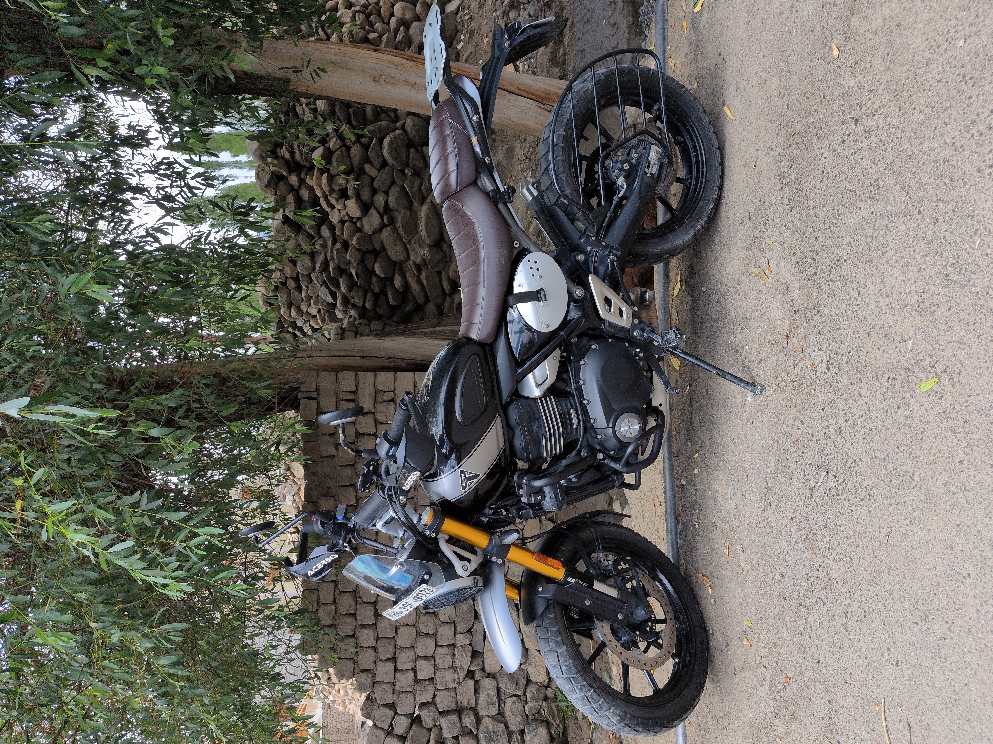 In the city of Leh, India, a black Triumph Scrambler motorcycle is parked on a dirt and gravel road, partially shaded by the branches of a mature tree. The motorcycle features a silver front fender, a clear windshield, a brown padded seat, and a silver rear luggage rack. The bike's license plate is visible and reads "WB-39F-8028". Behind the motorcycle, a dry-stone wall constructed from various-sized rocks forms a rustic backdrop. The surrounding environment is characterized by dry vegetation and a slightly overcast sky. The scene depicts a static moment, with no people or active events occurring.