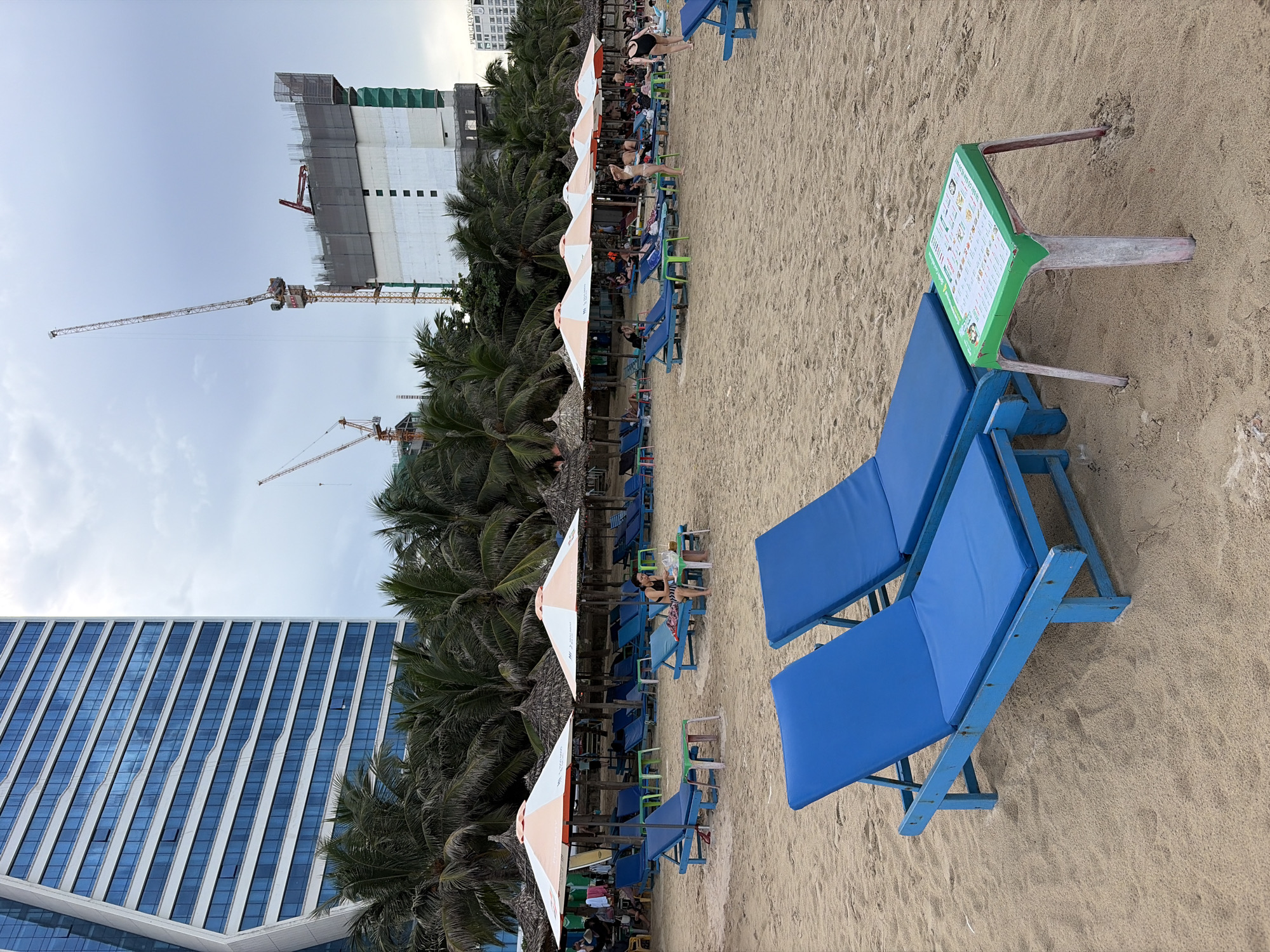 The image depicts a beach in Đà Nẵng, Vietnam, with a large modern building on the left and a construction site with cranes in the background. The foreground features two empty blue lounge chairs on the sand, with a small green table between them displaying a menu. Further back, a row of beach umbrellas and lounge chairs are set up, some occupied by people. Palm trees line the edge of the beach, separating it from the buildings and construction. The sky is overcast.