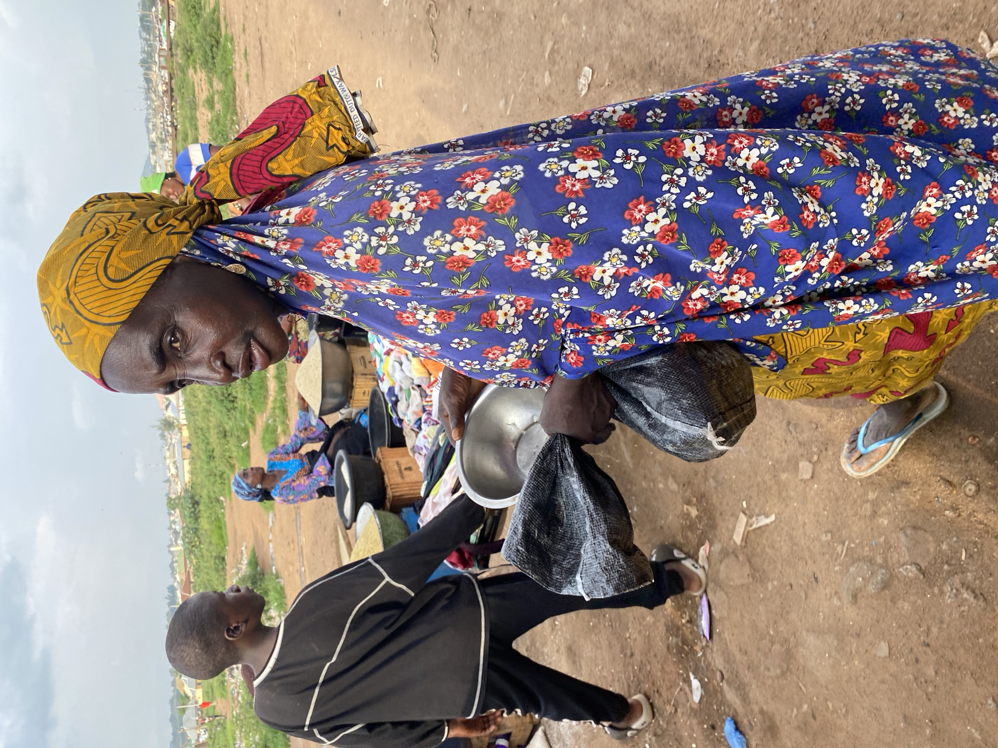 An outdoor market scene in Bukuru, Nigeria, shows a woman in the foreground wearing a blue dress with a floral pattern and a yellow headscarf. She holds a silver bowl and a black plastic bag. To her left, a young person wearing a black long-sleeved shirt and black pants is seen from behind, walking away from the camera. In the background, other people are visible, along with stalls selling goods, including grains in bowls. The ground is dusty and uneven, with some scattered litter. The sky is overcast. Buildings and vegetation are visible in the distance, indicating an urban or semi-urban environment.