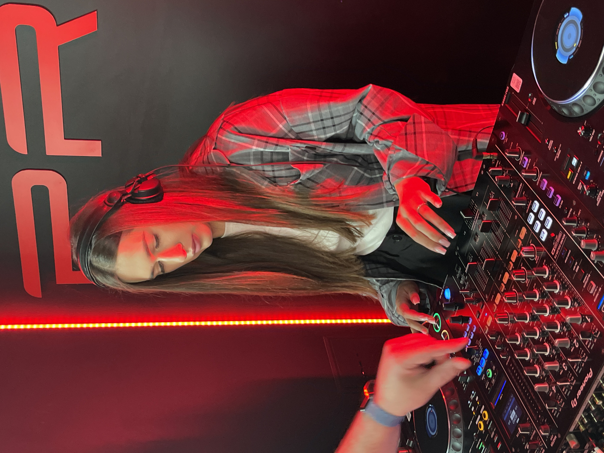 In a dark indoor setting, likely a club or studio in Kyiv, Ukraine, a young woman is engaged in DJing. She has long brown hair, wears headphones, a white top, a black vest, and a grey plaid oversized shirt. Her eyes are closed, suggesting deep concentration or immersion in the music. She stands in front of a professional DJ setup, which includes a Pioneer DJ mixer and at least one CDJ-3000 player. Another person's hand, partially visible in the foreground, is actively manipulating the controls on the mixer, indicated by a blue wristband.
The scene is dramatically lit by prominent red LED strip lights that run horizontally across the back wall, casting a strong red glow on the subjects and equipment. The background wall is dark grey or black, featuring large, stylized red letters, one clearly an "R" and another partially visible. The DJ equipment also has various illuminated buttons and displays, adding to the vibrant atmosphere. The overall impression is one of an energetic music performance or recording session taking place at night.