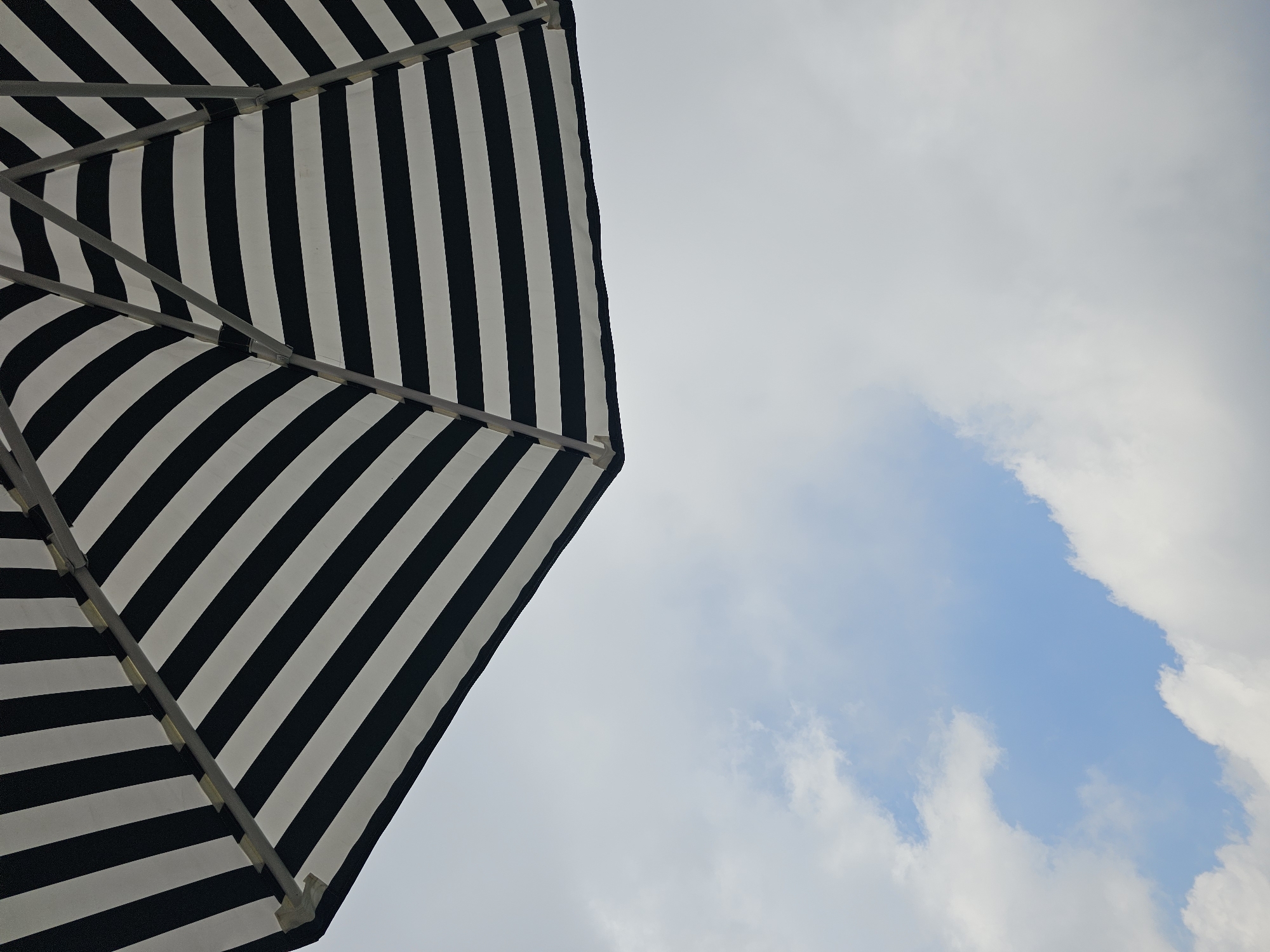 An under-view of a black and white striped umbrella is captured against a cloudy sky. The umbrella's fabric is taut, with a pattern of alternating black and white stripes. The metal framework of the umbrella is visible, with silver poles radiating from a central point. The umbrella occupies the upper portion of the image. The lower portion of the image shows a sky with soft, white clouds and patches of blue. The scene does not contain any people or specific details indicating a location in Kintamani, Indonesia.