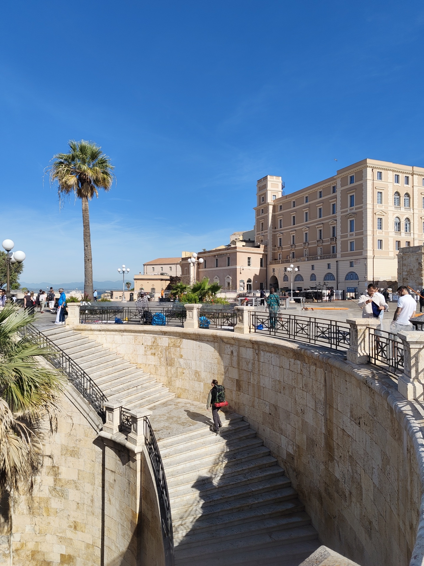The image depicts a sunny daytime scene in Cagliari, Italy, featuring a multi-tiered staircase made of light-colored stone with black metal railings. A lone person wearing a dark jacket, backpack, and carrying a red bag is descending the stairs. The stairs are surrounded by a curved stone wall.
Above the staircase, a public space is visible, with several people standing and walking along a balustrade with decorative metalwork. A tall palm tree is visible on the left side of the image. In the background, there is a large, light-colored building with multiple windows and a tower-like structure. The sky is clear and blue. Several other people are milling about, including some near railings overlooking a lower area. There also seem to be some preparations for an event in the center.